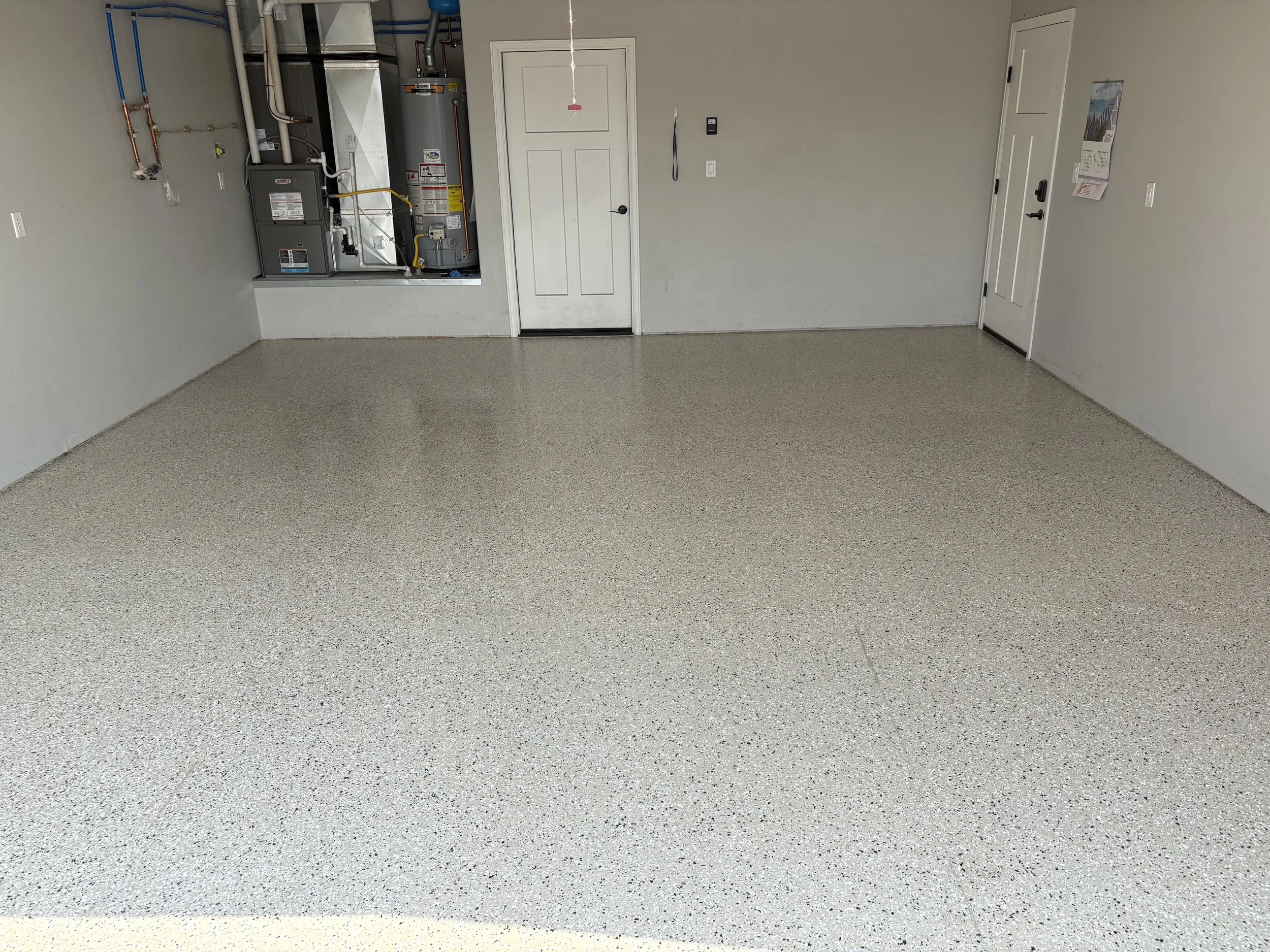 Empty garage with light-colored speckled floor, plain walls, a white door, a water heater and furnace on the left, and a calendar hanging on the right wall.