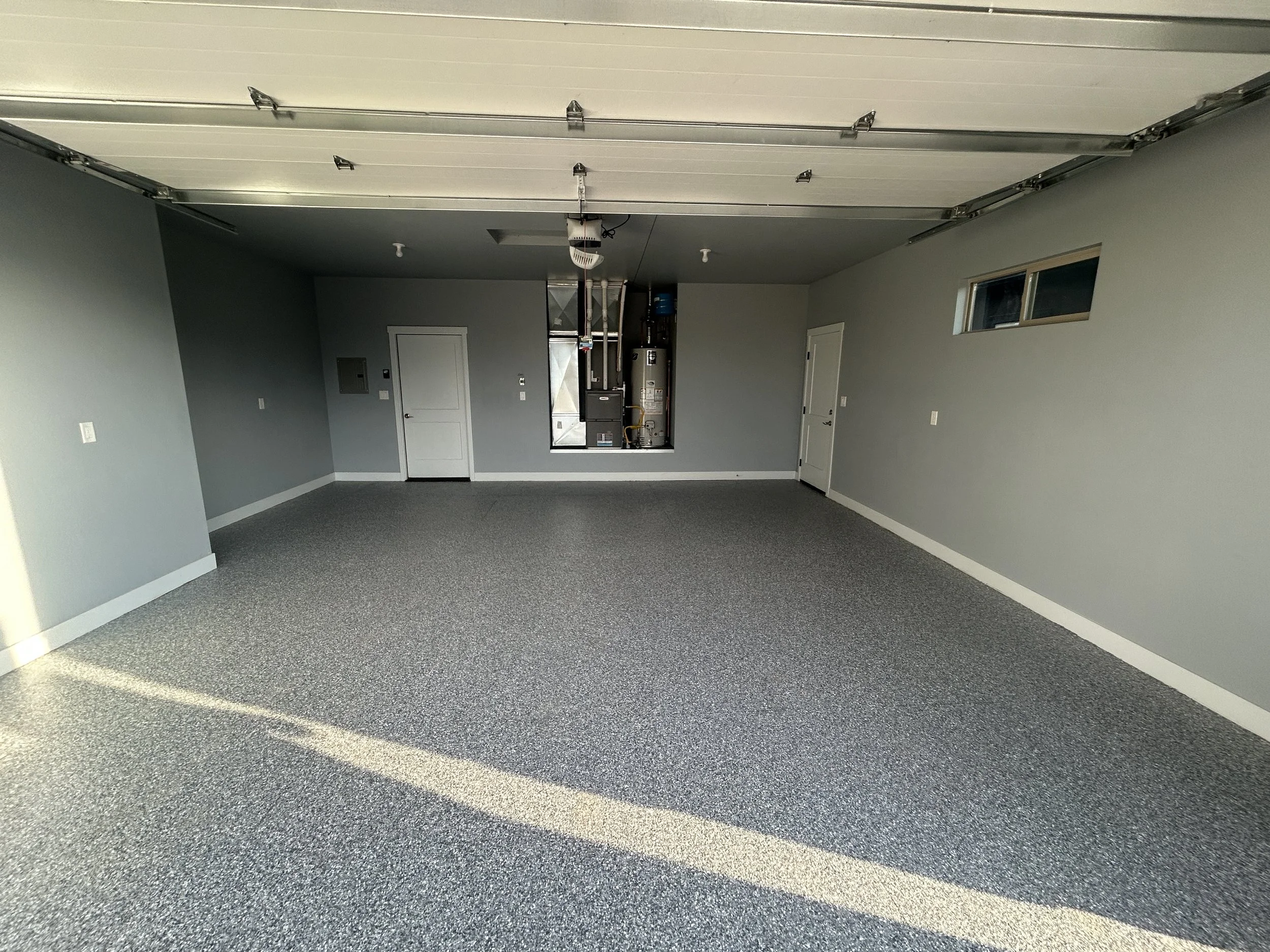 Empty garage with gray walls, gray carpet, and a white garage door. There is a small window on the right wall and an open access panel for utilities on the left wall. Two closed white doors are at the back.
