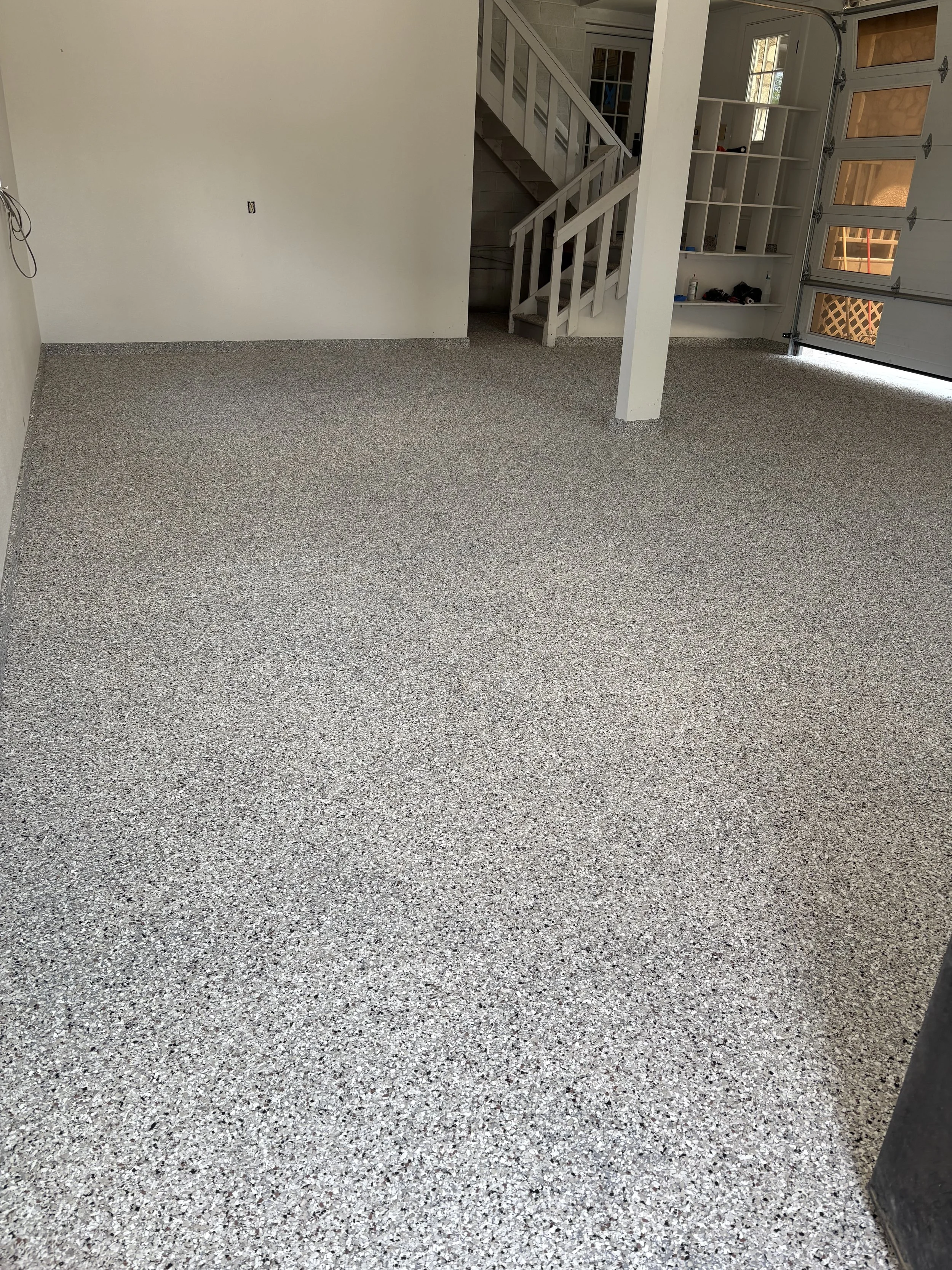 Interior of a garage with a speckled epoxy floor, white walls, a staircase leading upstairs, and a closed garage door with windows.
