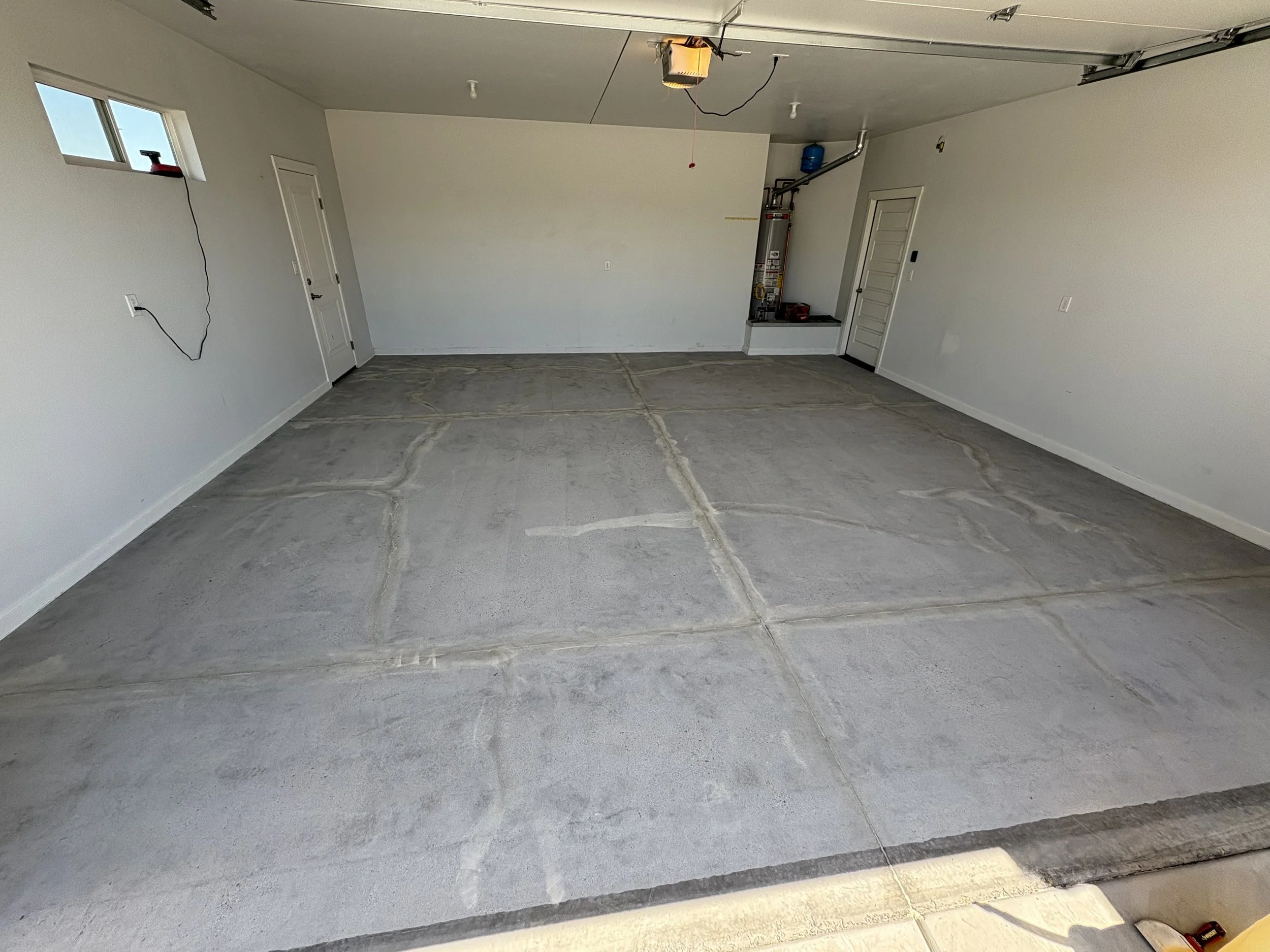 Empty garage with plain white walls, a concrete floor, and a garage door open. There is a small window and a closed door inside. Garage equipment includes a water heater, a blue water pressure tank, and some tools on a small platform.