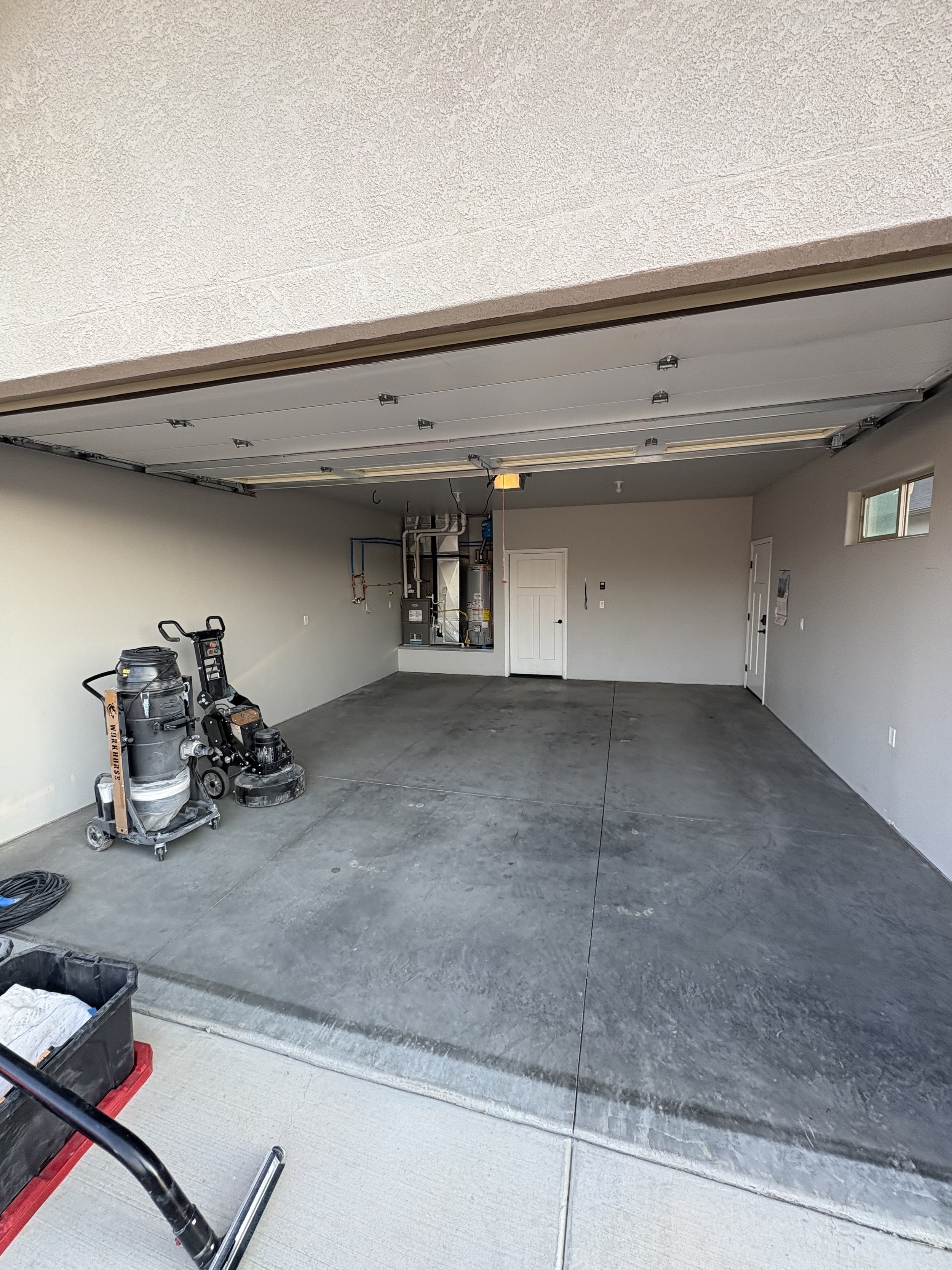 View of a partially completed garage with a concrete floor, walls, and some tools and equipment present. The garage door is open, showing the interior space.