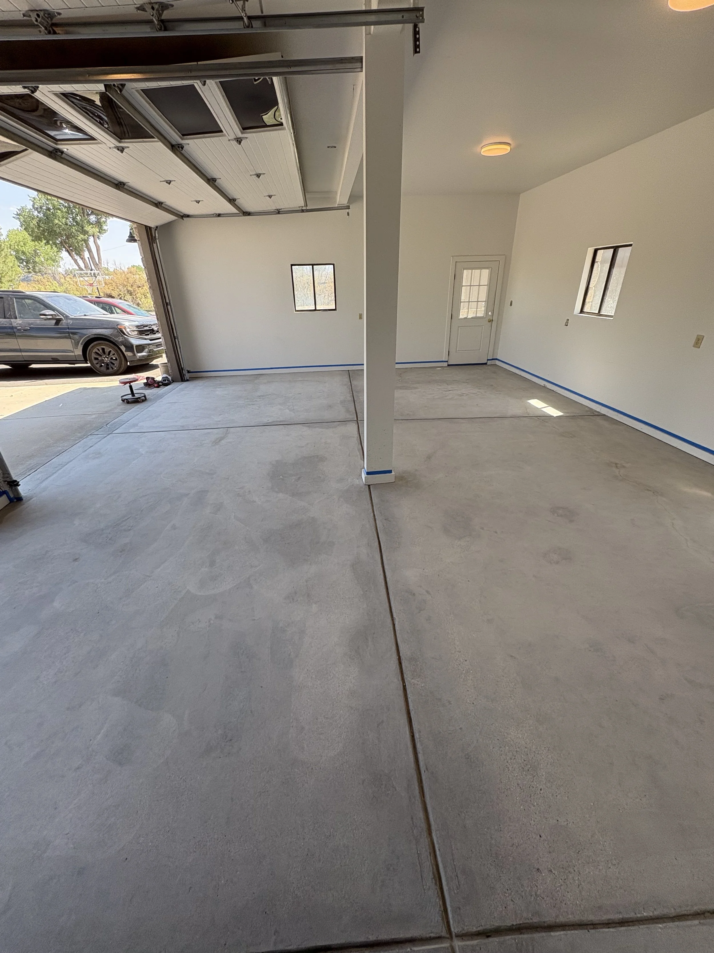 An unfinished garage with concrete floor, white walls, and an open garage door. Outside, there are parked cars, trees, and a basketball hoop visible.