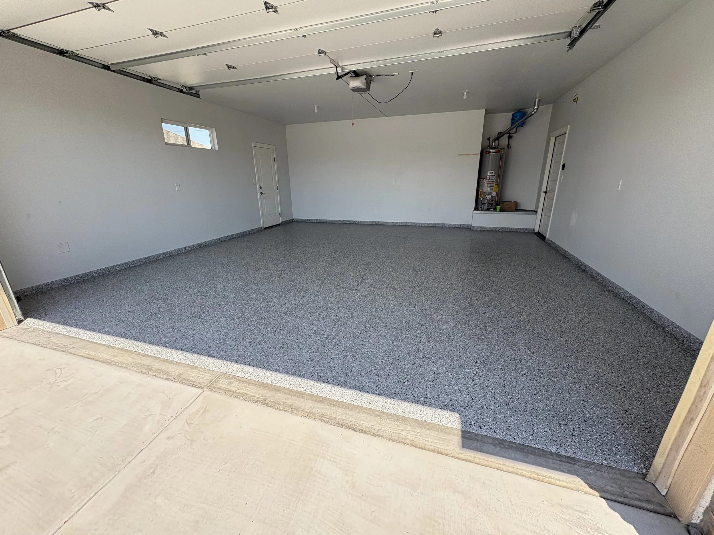 Empty garage with a grey speckled epoxy floor, white walls, a small window, and a water heater in the corner. Garage door is open, showing the metal tracks and garage door opener.