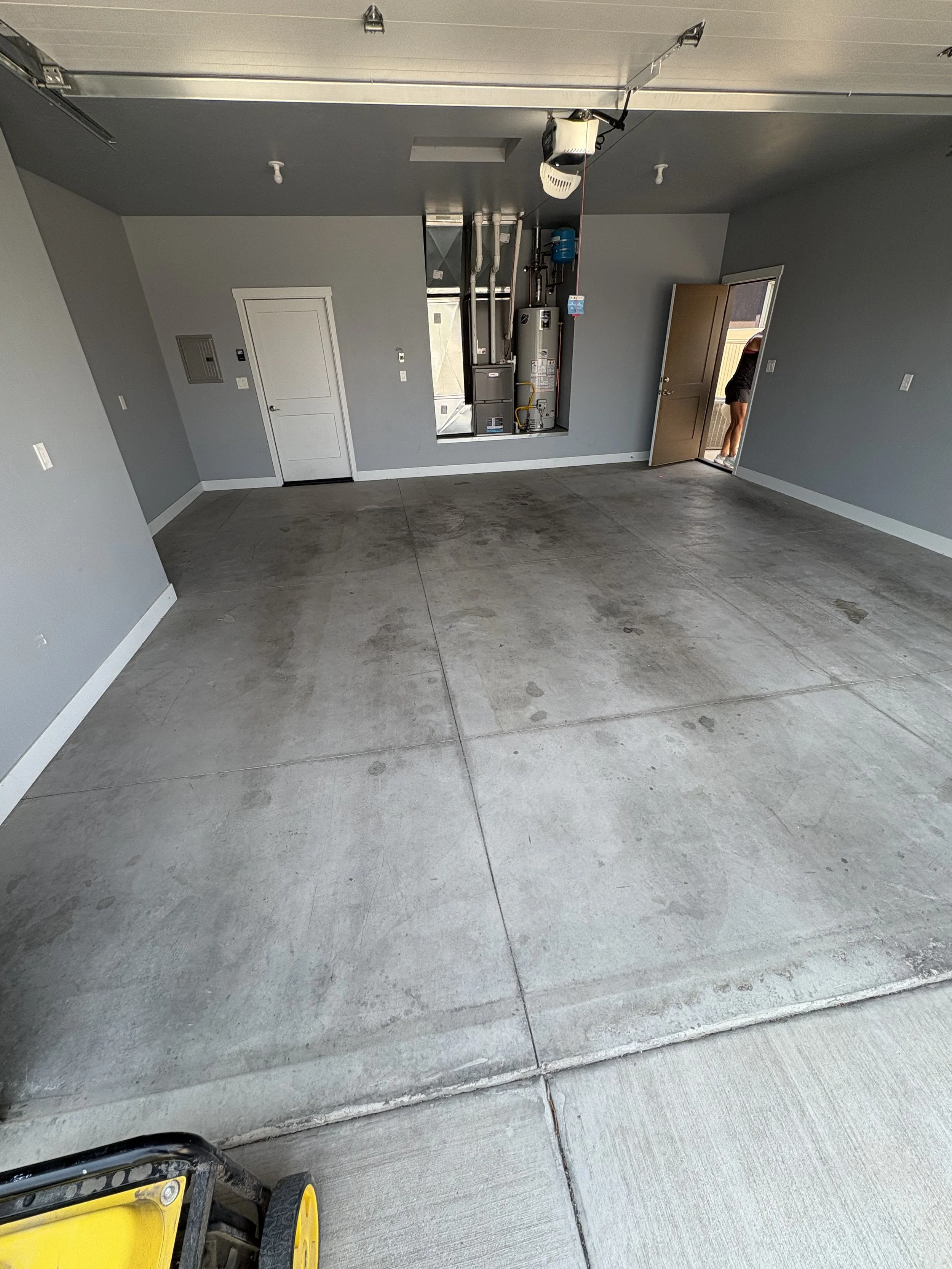 Empty garage with a concrete floor, gray walls, a water heater and utility box on the wall, and an open door with a person partially visible outside.