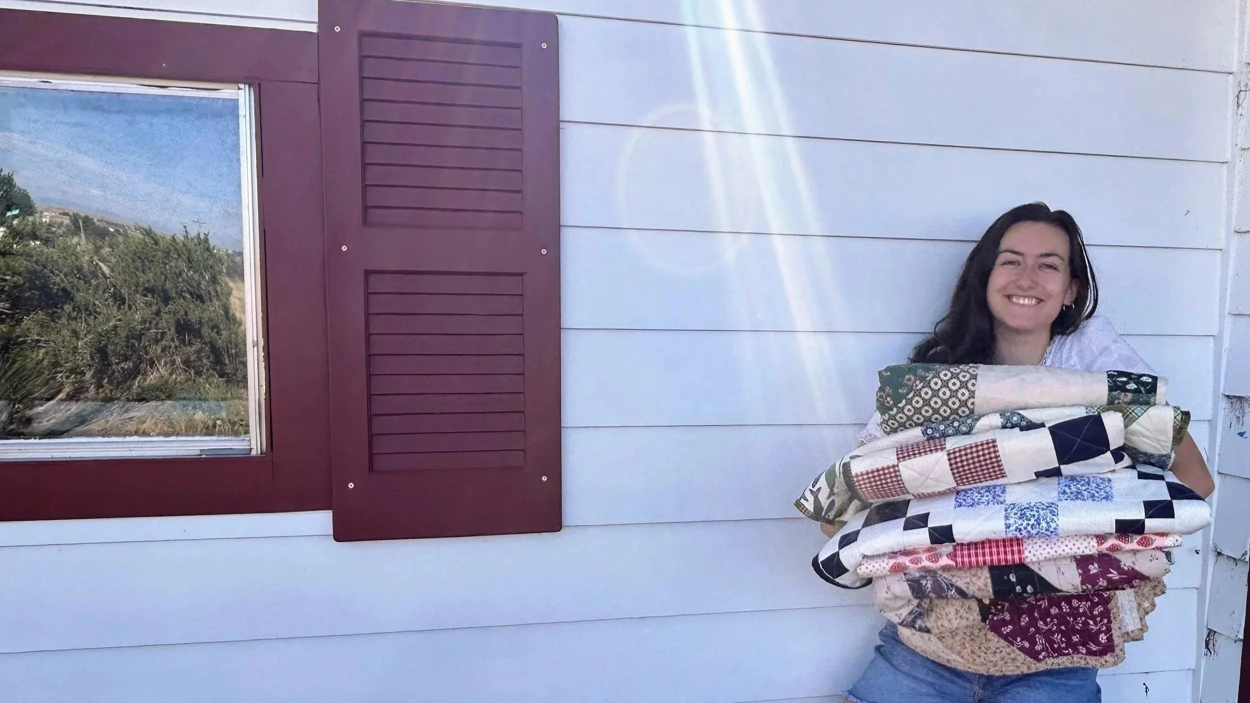 A woman smiling and holding a large stack of folded quilts outside a house with white siding, a window with purple shutters, and a view of trees and hills in the background.