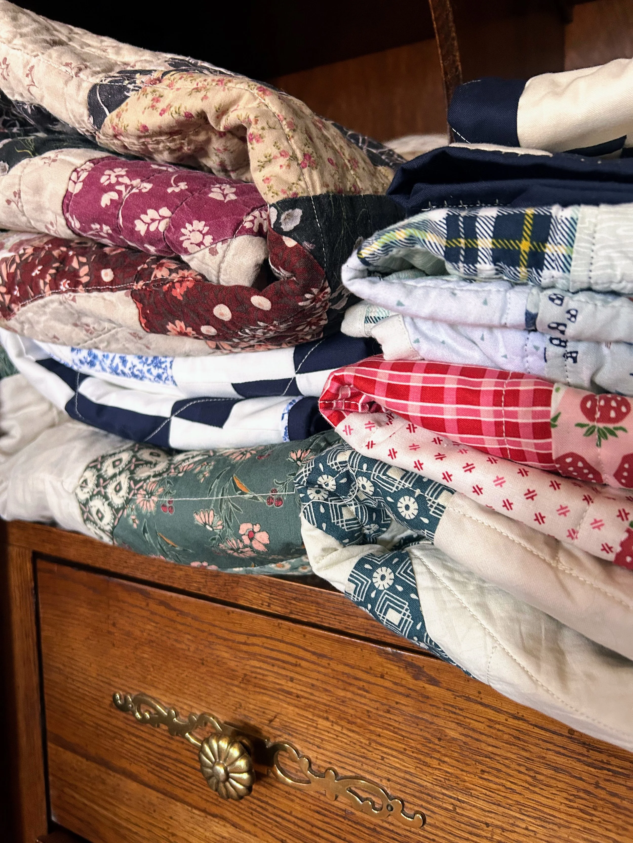 Stacked folded quilted and patterned fabric pieces on a wooden furniture drawer with ornate metal handle.