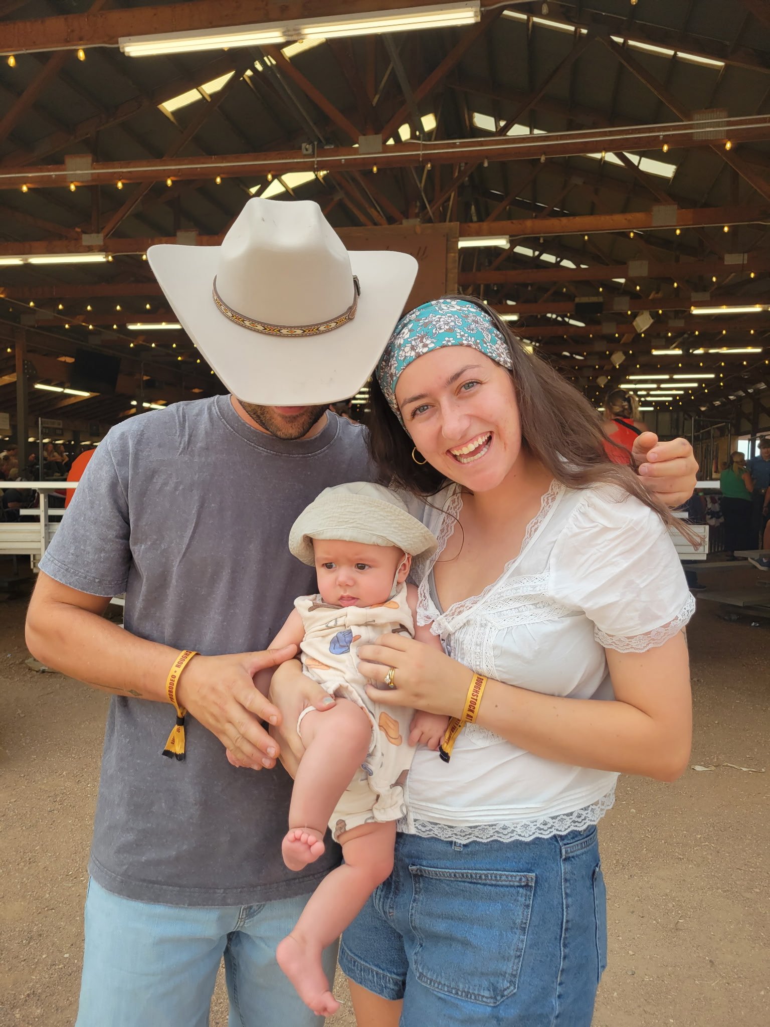 Smiling woman holding a baby girl with a man in a white cowboy hat standing beside them indoors, with some people and a barn-like structure in the background.
