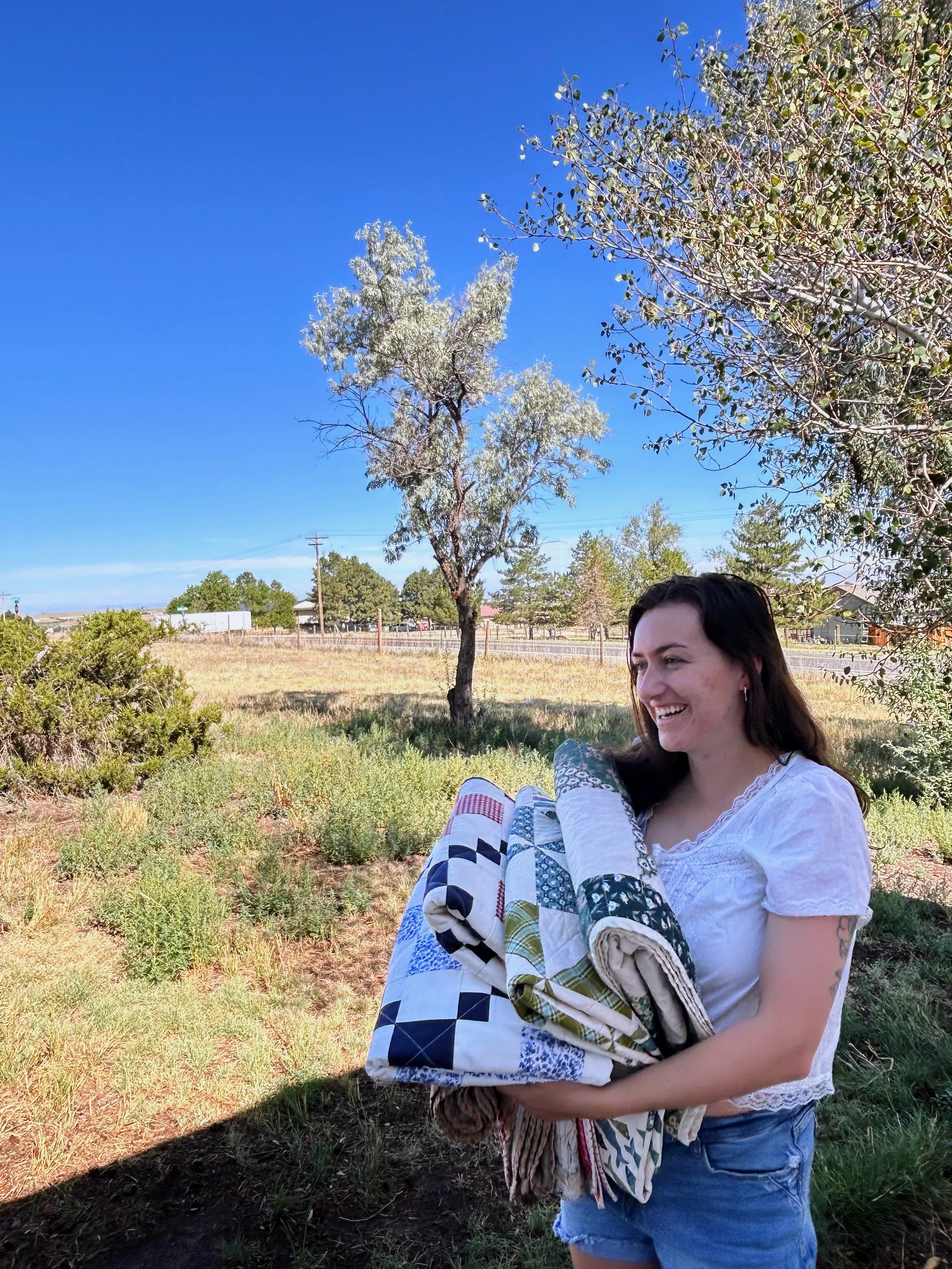 A woman with dark hair and tattoos on her arm smiling while holding and hugging a quilt or blanket outdoors in a grassy area with trees and blue sky in the background.
