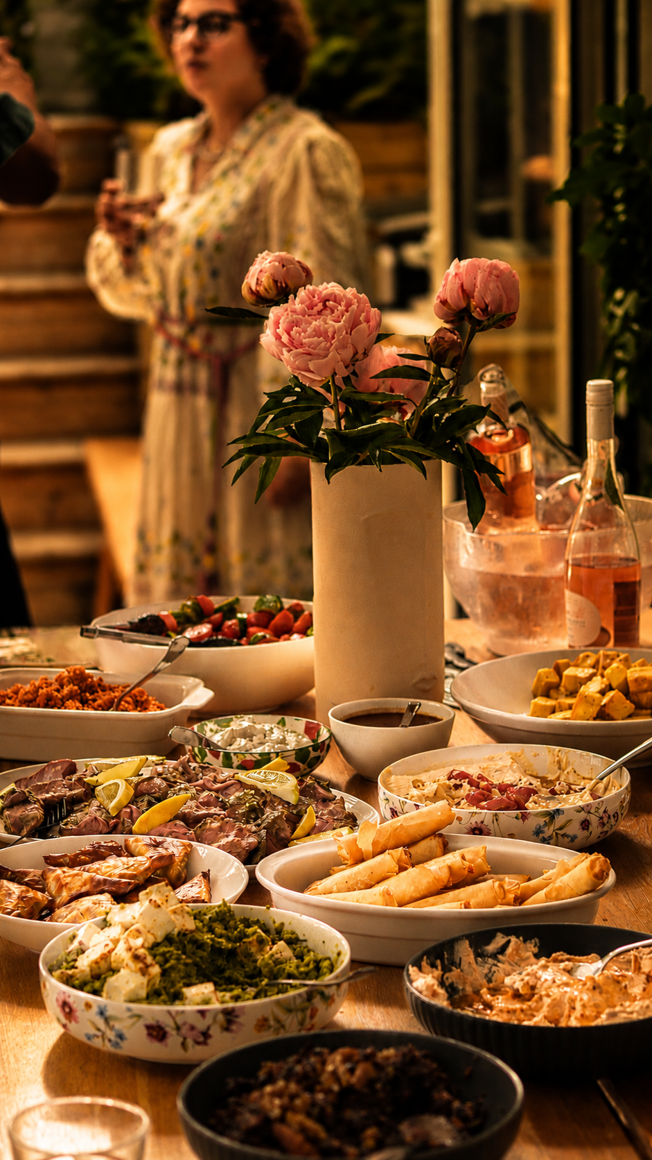 A table set with various dishes and a vase of pink peonies, with a woman in the background holding a glass, at an outdoor gathering during evening.