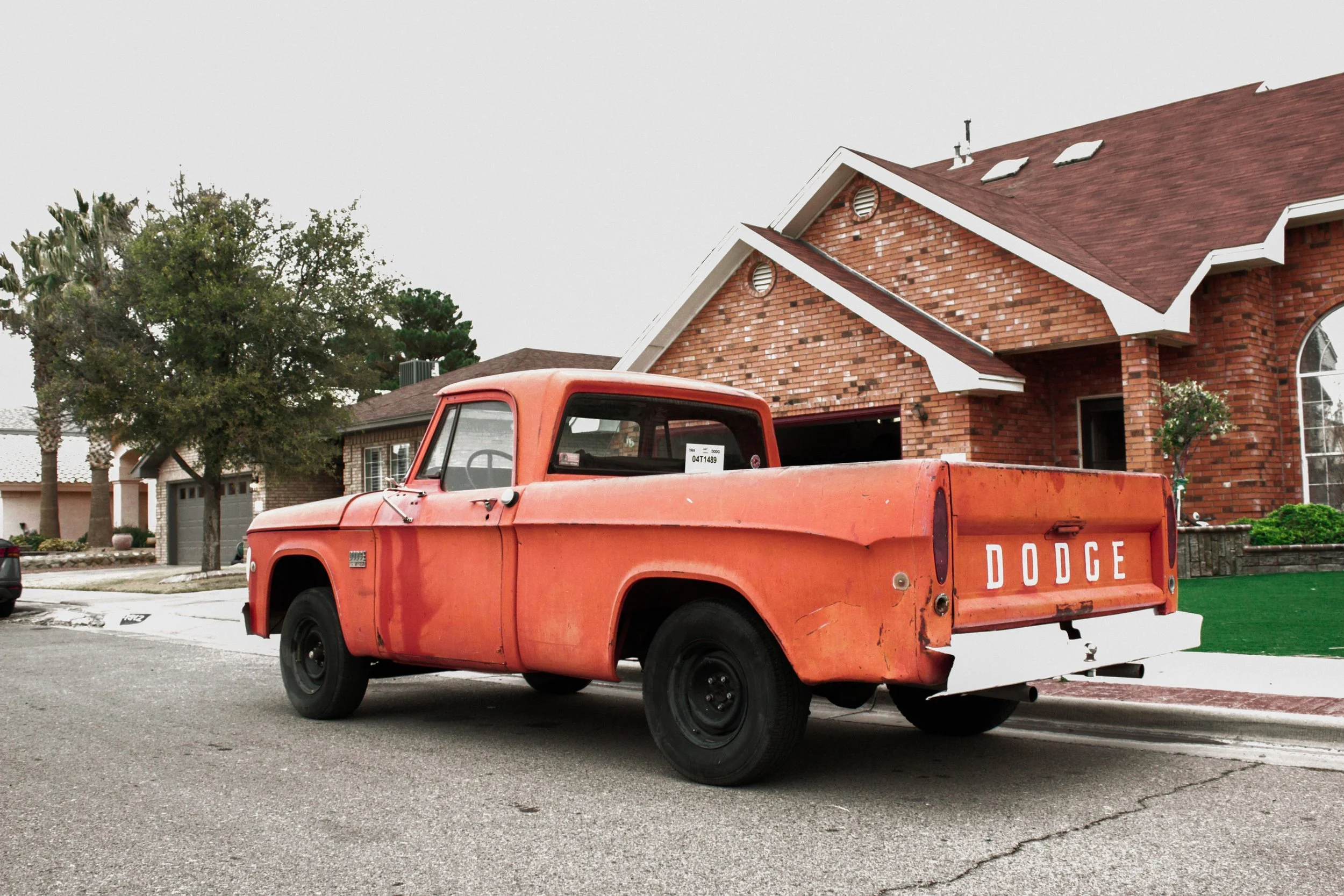 An old red Dodge pickup truck parked in front of a brick house with a large arched window, surrounded by trees and neighbor houses.