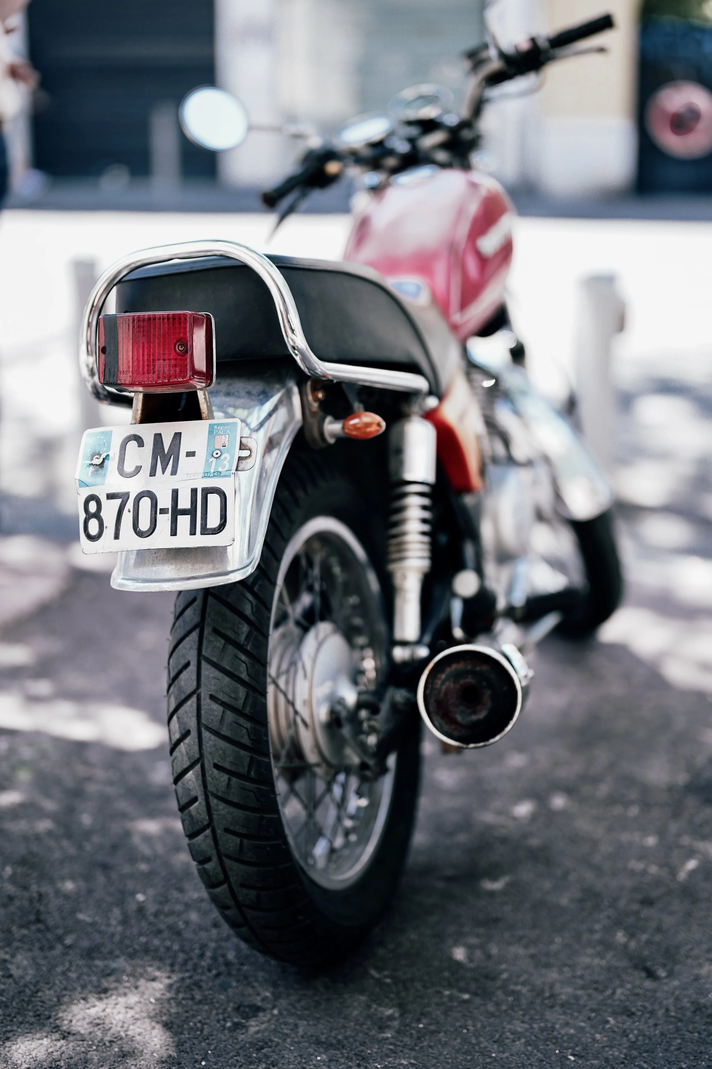 Close-up of a motorcycle's rear wheel, license plate, tail light, and exhaust pipe, parked on a paved surface with shadows.
