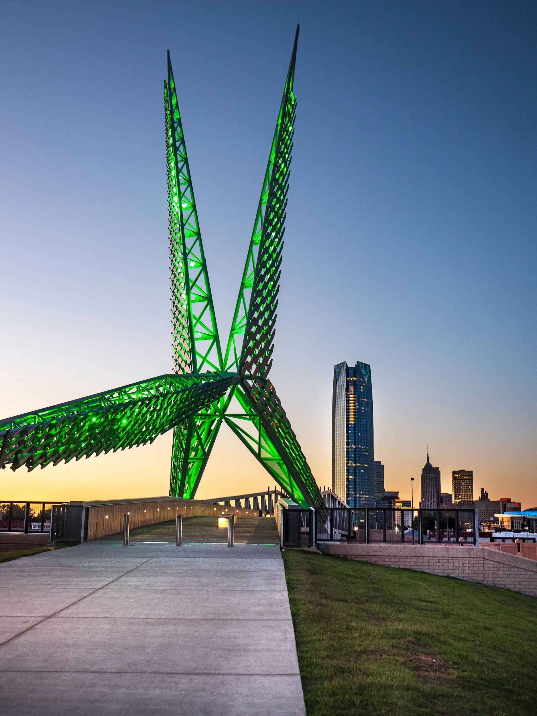 A modern green-lit sculpture resembling a star with curved, pointed arms, situated in an urban park with city skyscrapers in the background at sunset.