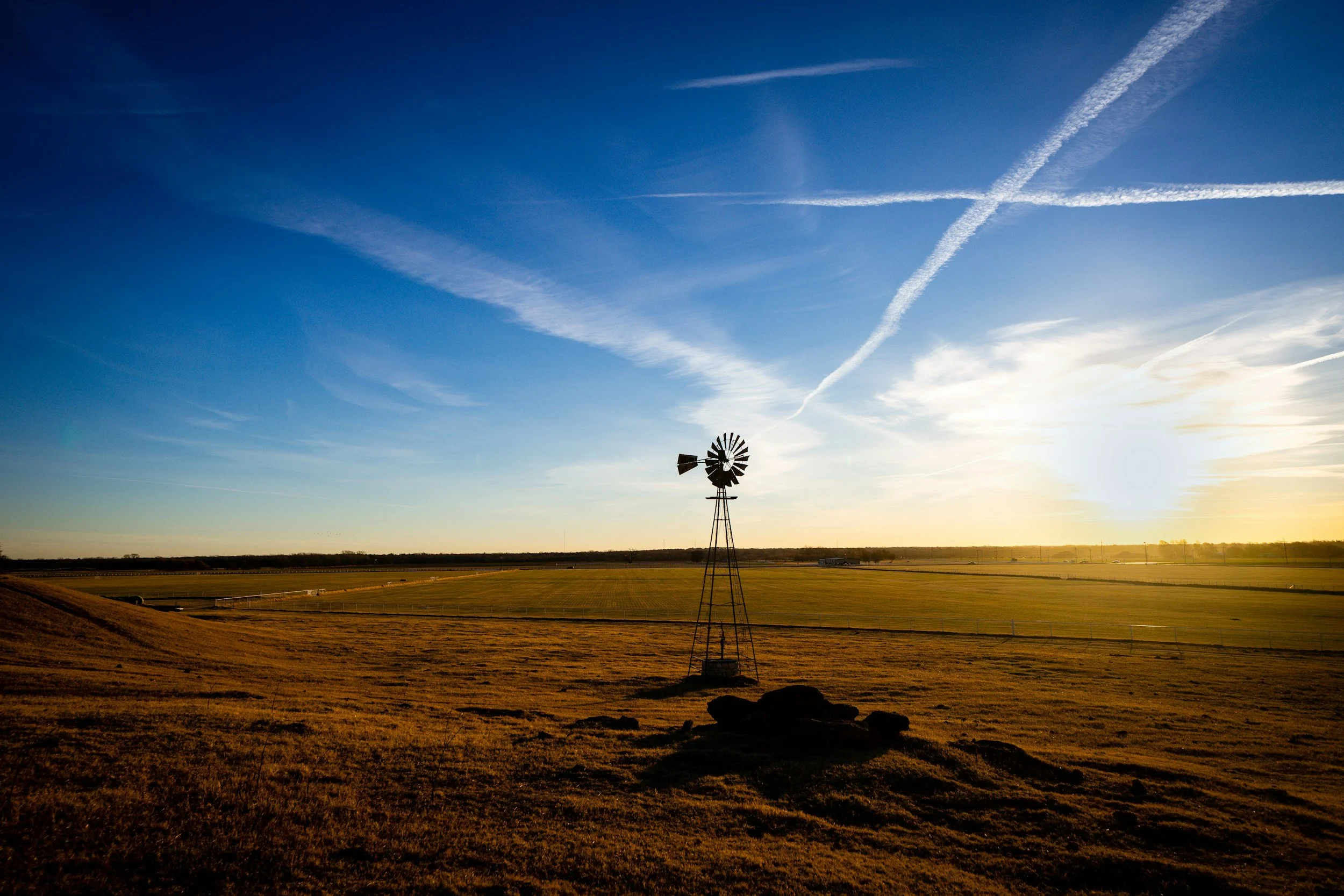 A rural landscape at sunset featuring a windmill in the foreground, open fields, and a sky with contrails and scattered clouds.