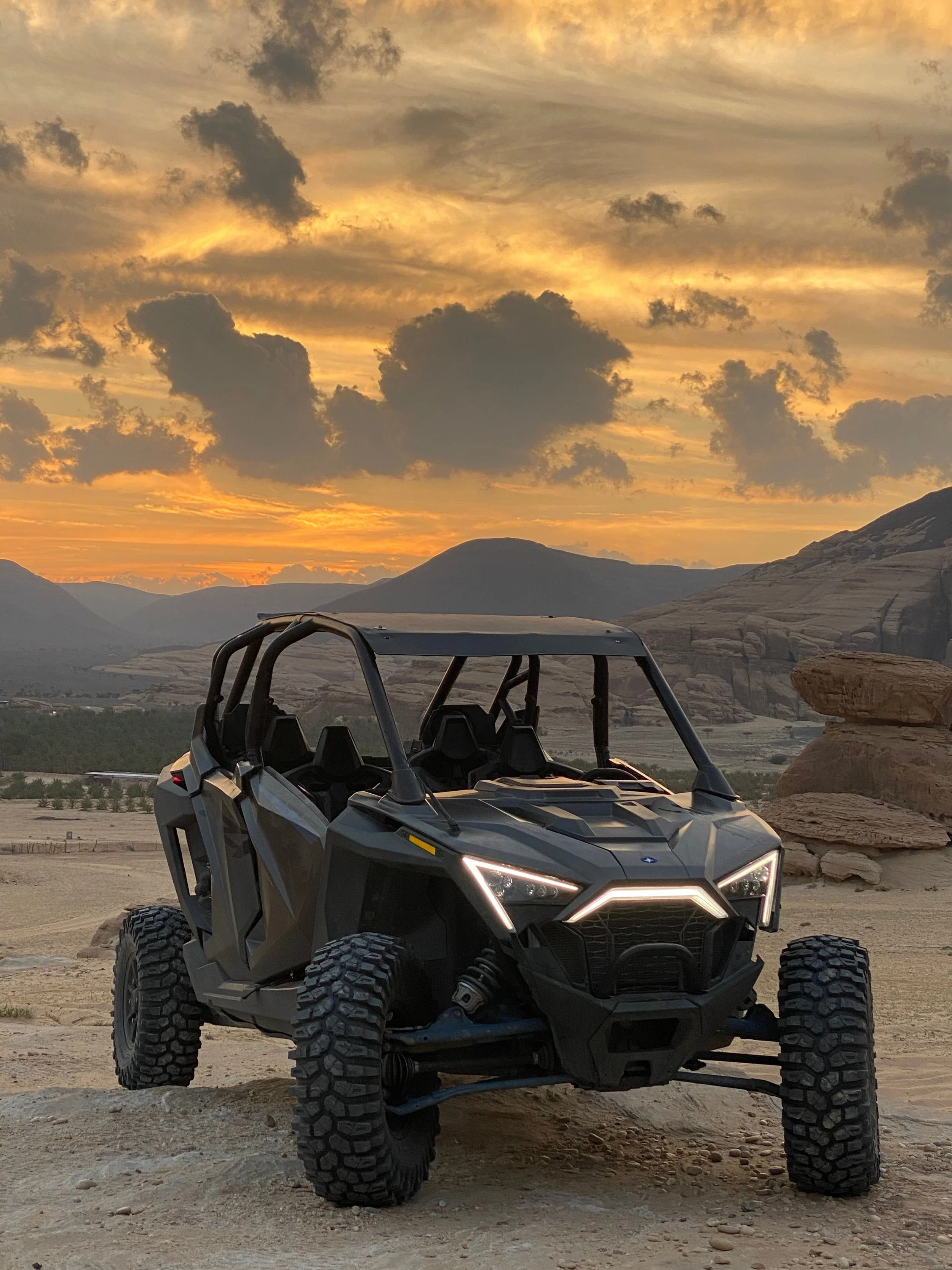 A black off-road utility vehicle on a desert landscape during sunset with mountains and clouds in the background.