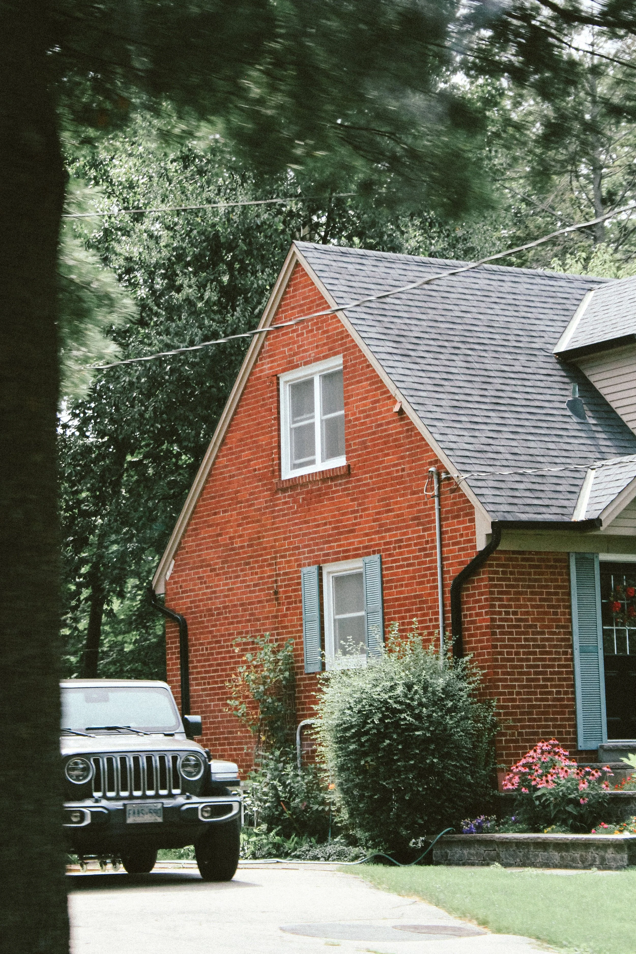 A red brick house with blue shutters, a gray shingle roof, and a white-framed window sits behind a driveway and lush bushes. A white luxury vehicle is parked in the driveway, and colorful flowers and green grass are in the front yard. Tall trees surround the house, and power lines are visible overhead.