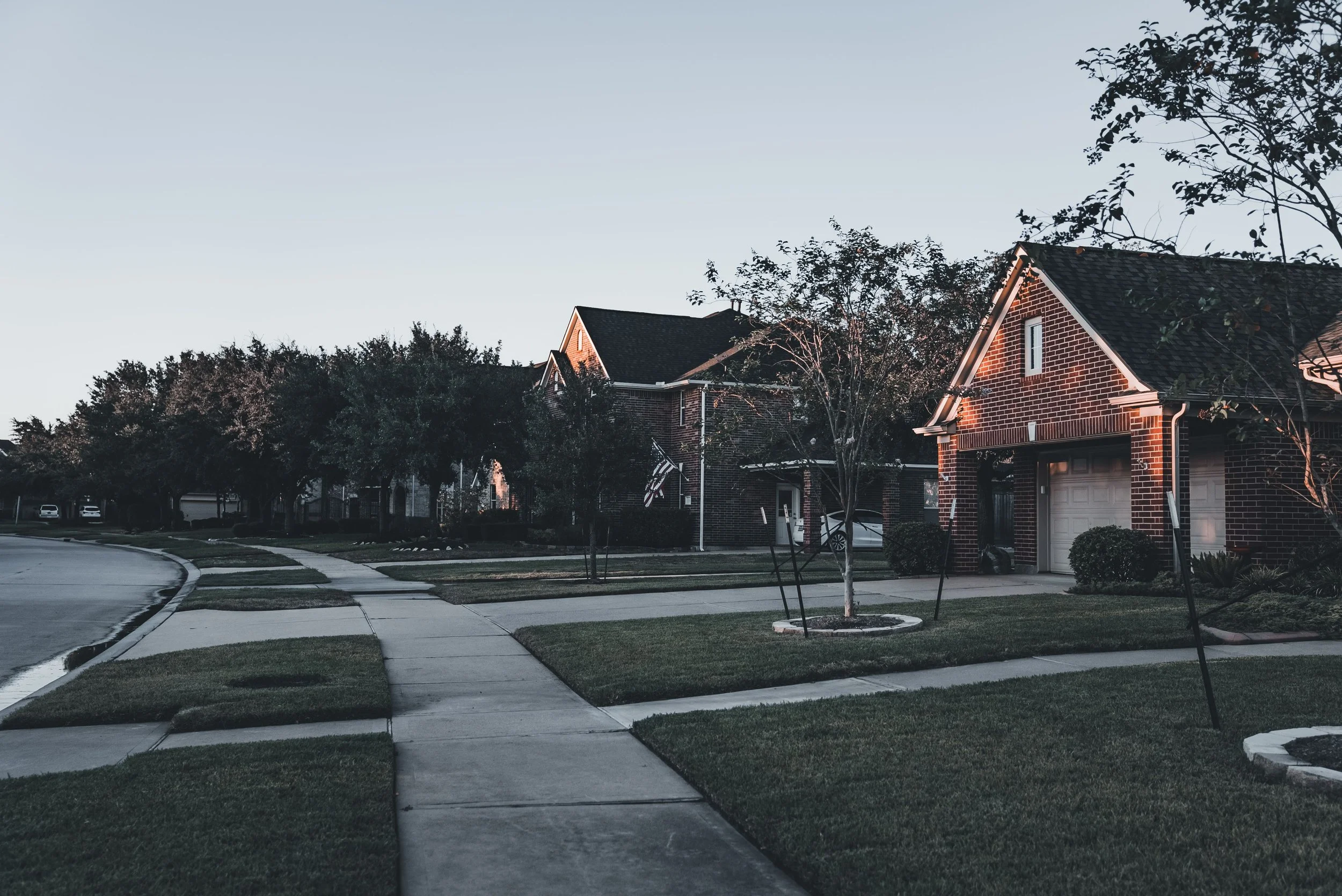 A suburban neighborhood street at dusk with brick houses, trees, a curved sidewalk, and neatly maintained lawns.