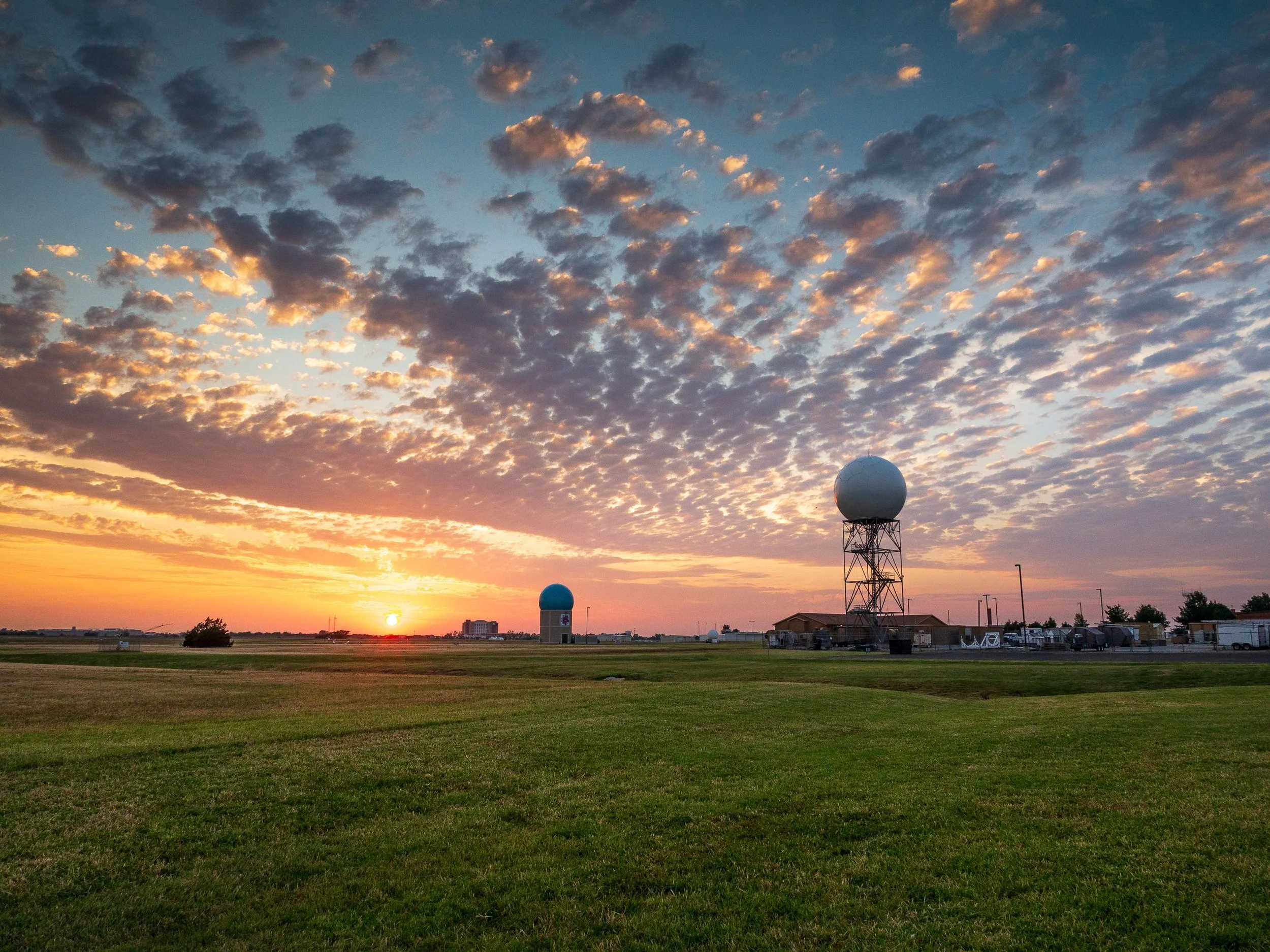 A sunset over a flat landscape with grass and a few trees, featuring radio or radar towers with spherical tops and a clear sky with scattered clouds.