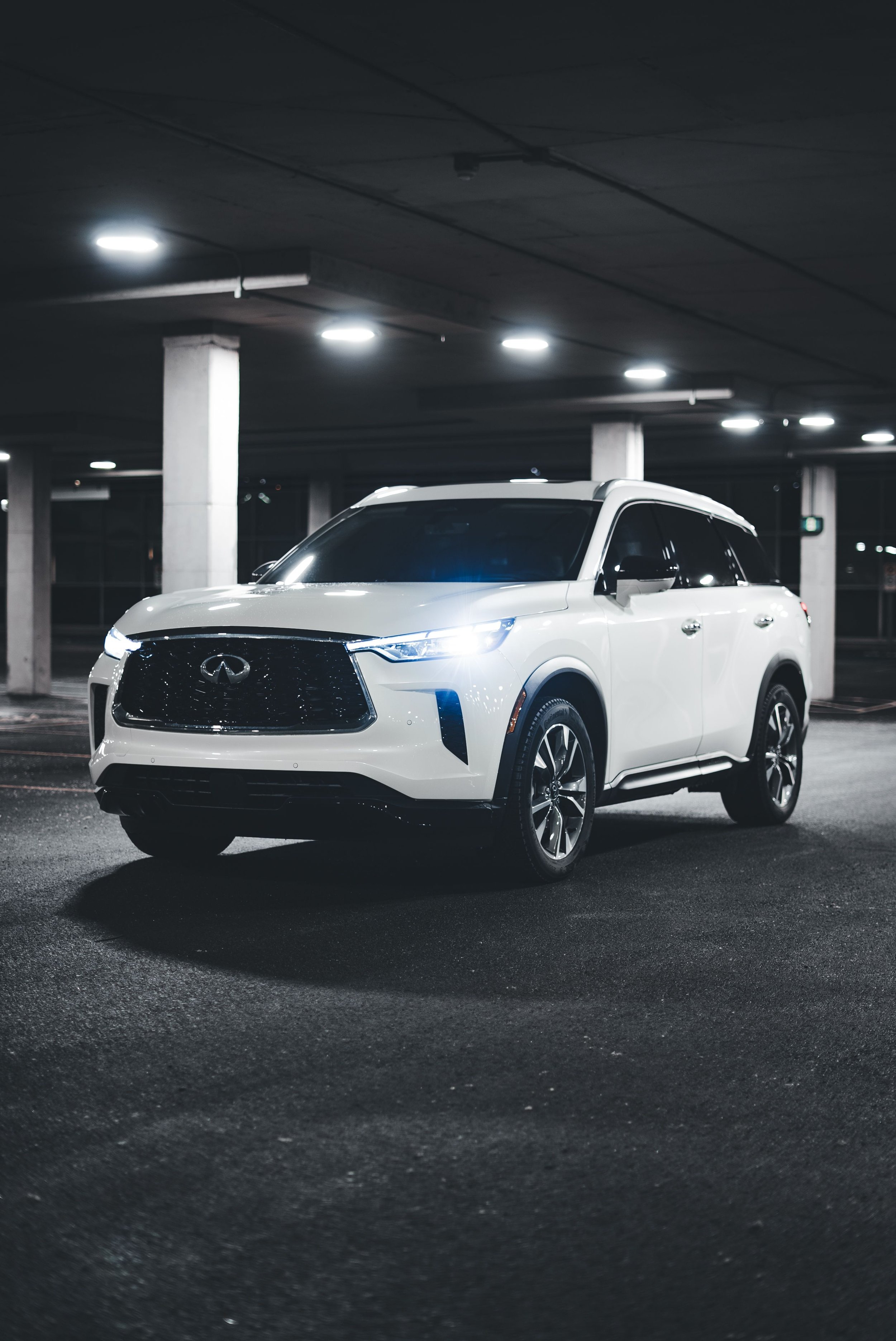 A white Infiniti SUV parked in an underground parking garage with overhead lights illuminating the space.