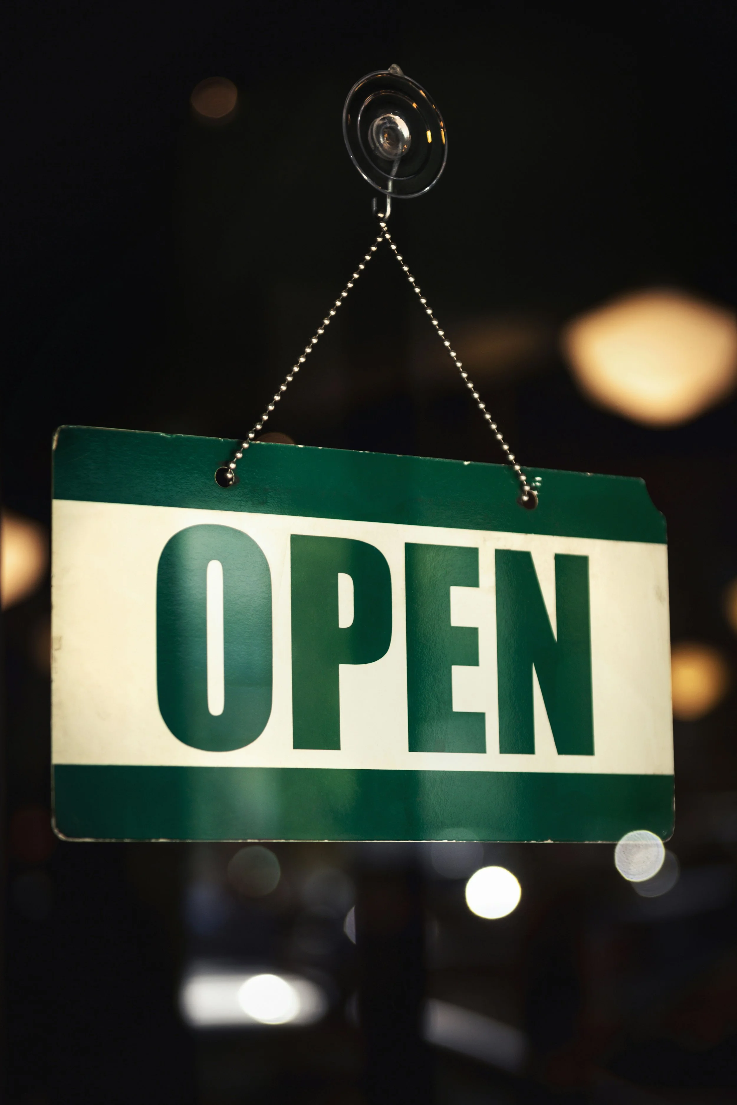A green and white sign with the word "OPEN" hanging from a chain and wall hook against a dark background, illuminated by dim lighting.