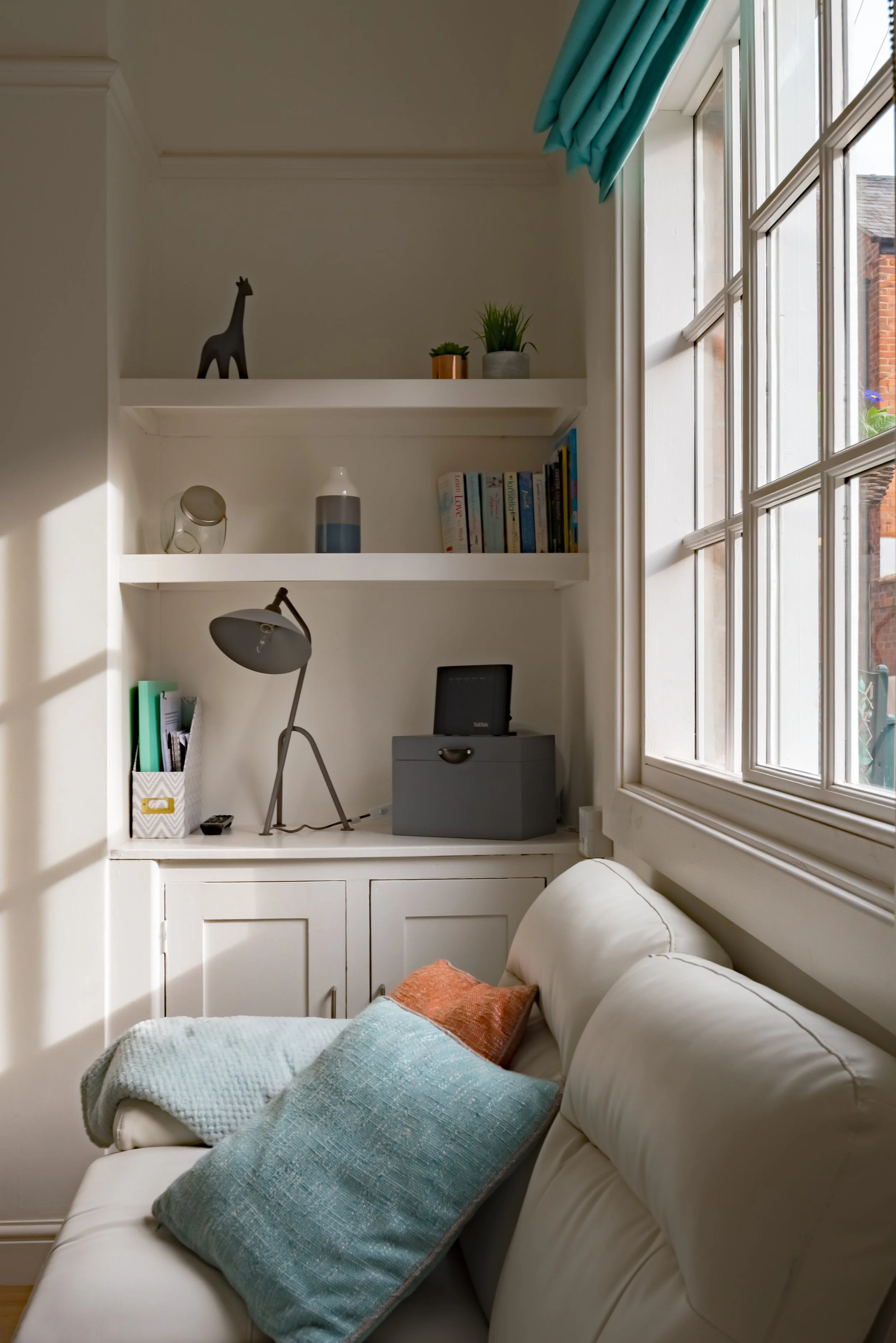 A cozy living room corner with a white sofa adorned with large teal and orange pillows. A white built-in shelf above holds decorative items, plants, and books. Sunlight streams through a large window with a blue curtain.