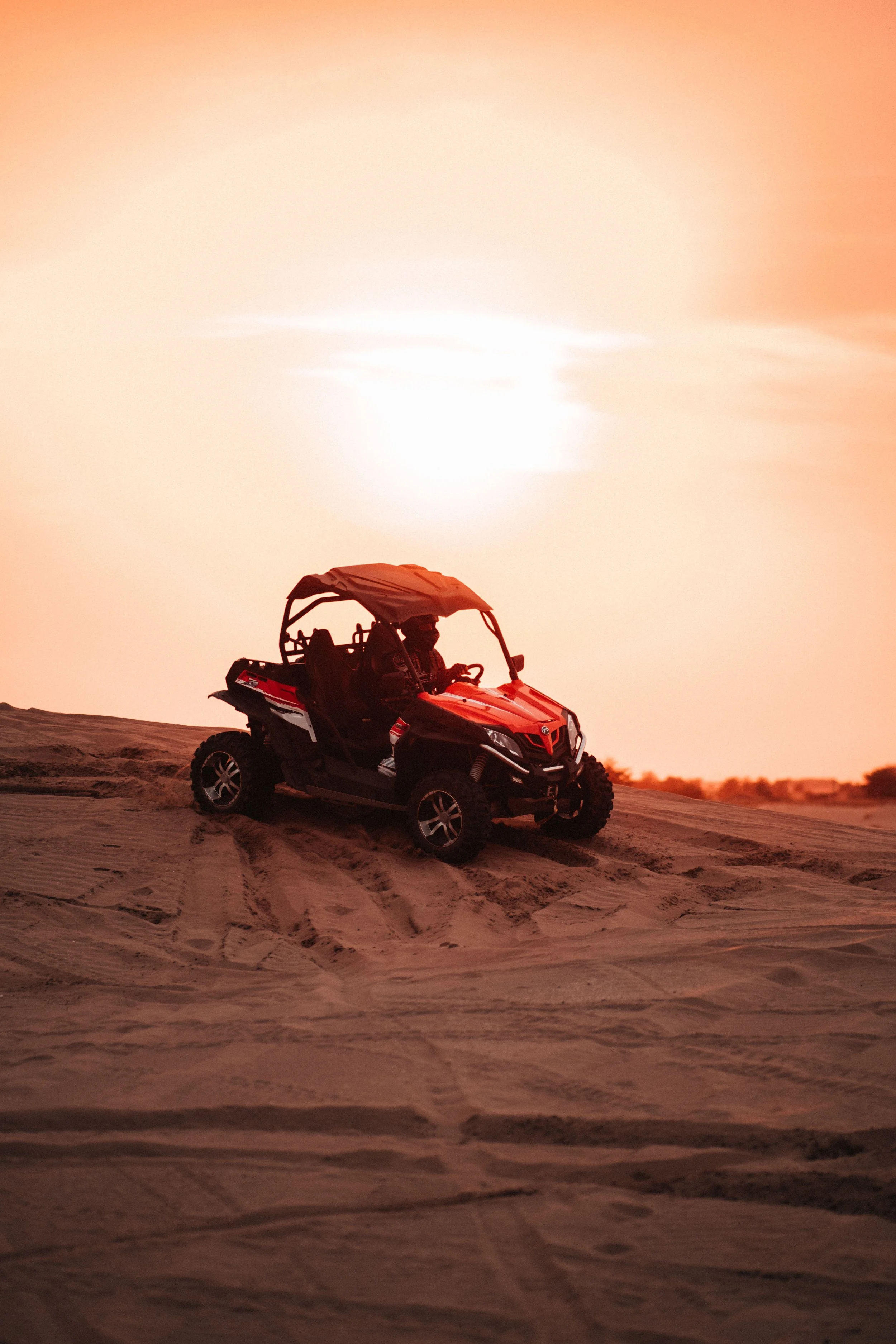 A small off-road vehicle, possibly a dune buggy, sitting on sand dunes at sunset with the sun low on the horizon.