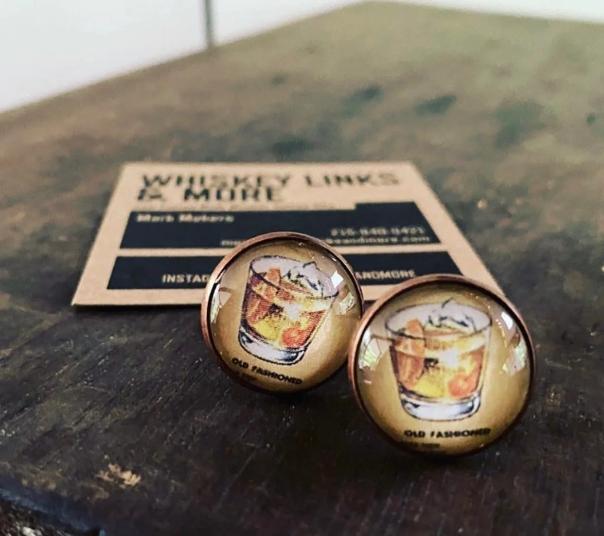 A pair of round cufflinks with a beer mug illustration and the text 'Old Flavour' on a dark wooden surface, with a minimalist card in the background.