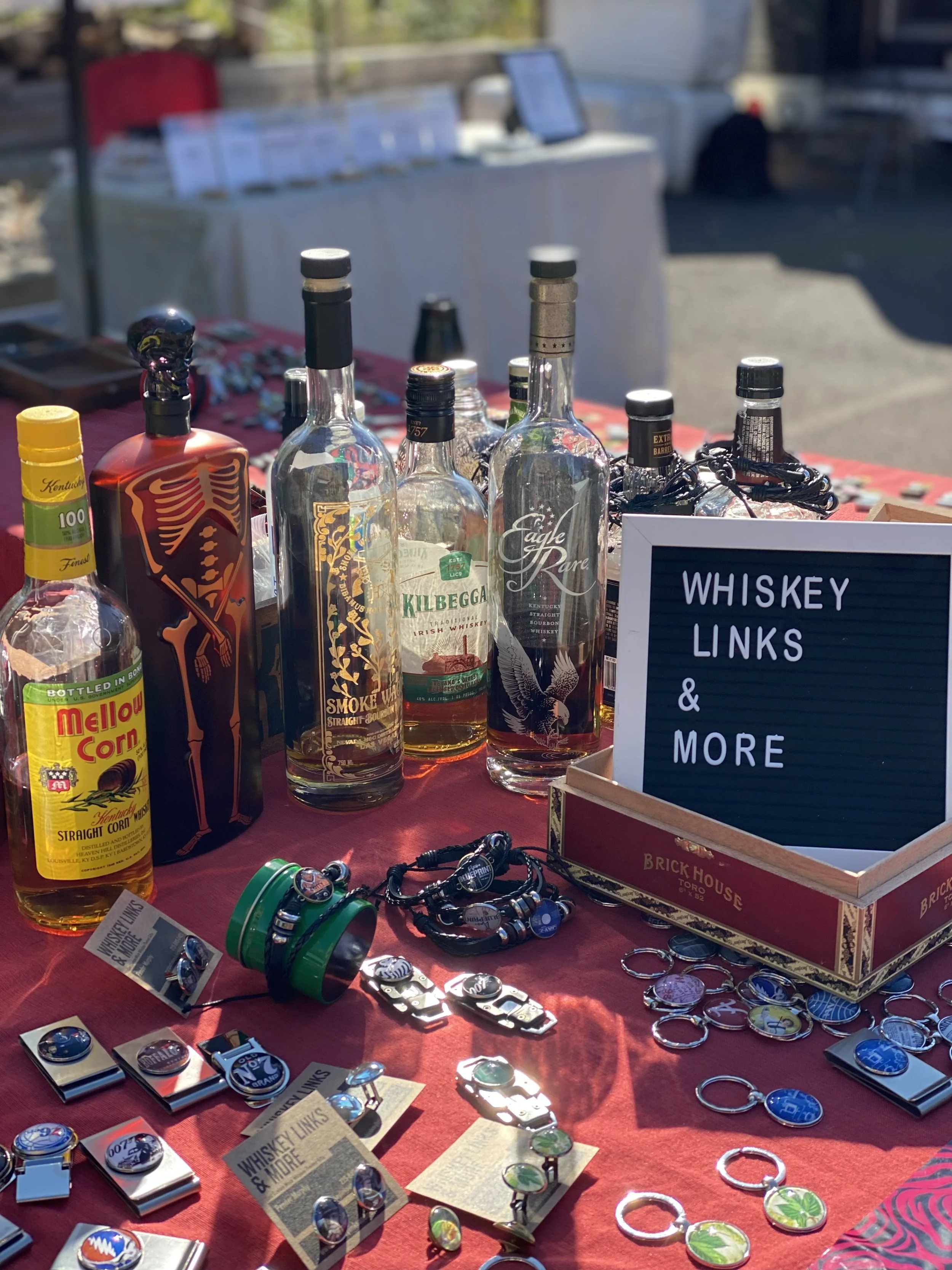 Display of whiskey bottles, keychains, and bottle openers on a red tablecloth at an outdoor event; sign reads 'Whiskey Links & More'.