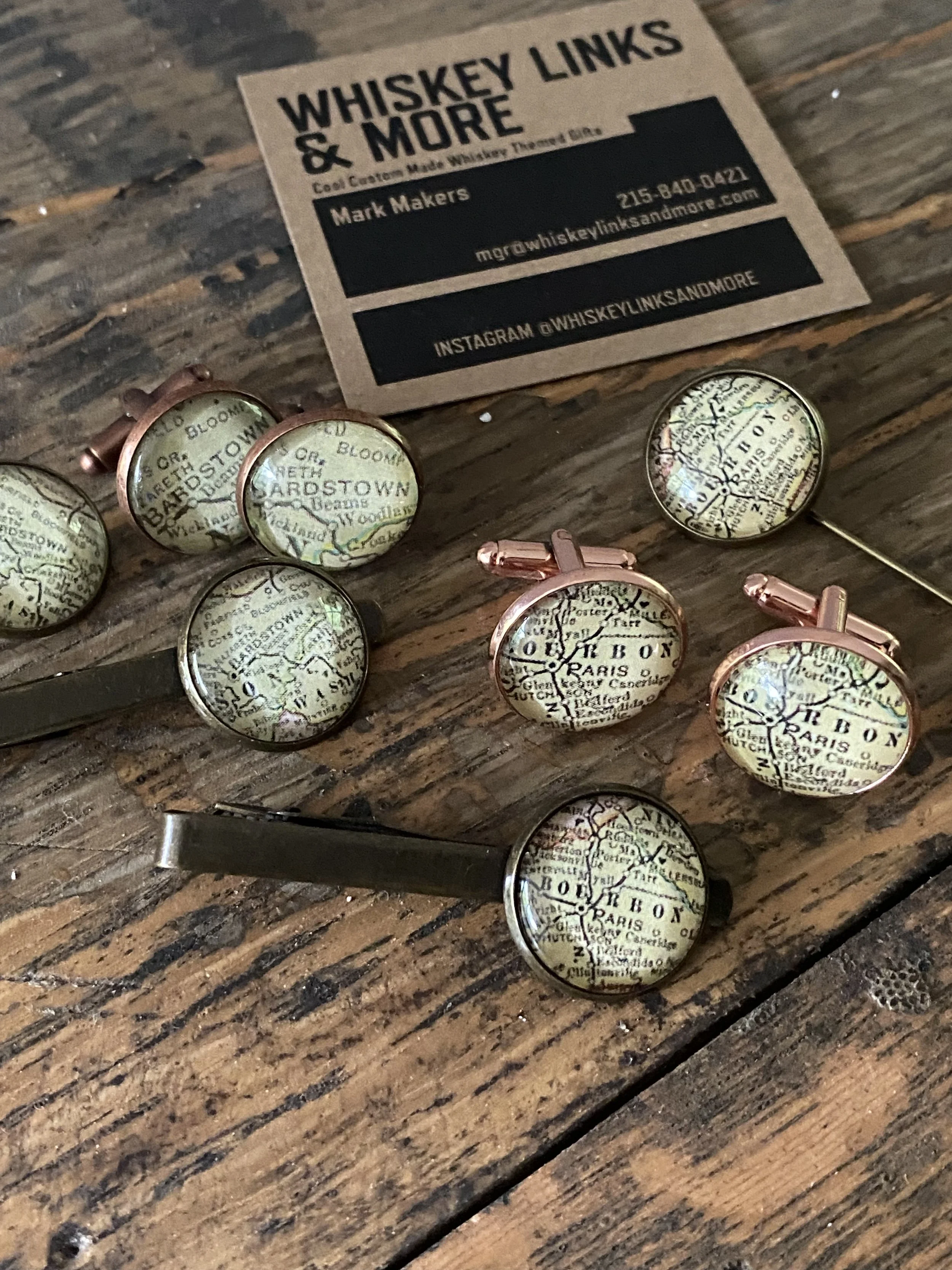 A business card for Whiskey Links & More featuring contact information and social media handle, placed on a wooden surface. Several vintage-style cufflinks with map designs are also on the surface.