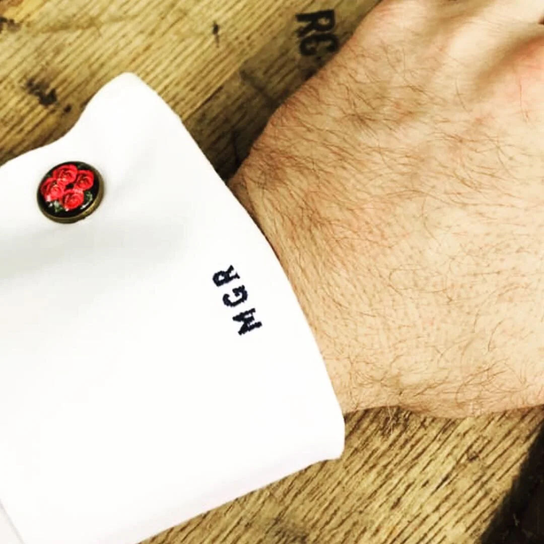 Close-up of a person's wrist with a white shirt cuff and black embroidered initials 'HCR', showing a small tattoo of a letter 'L' on the wrist, resting on a wooden surface.