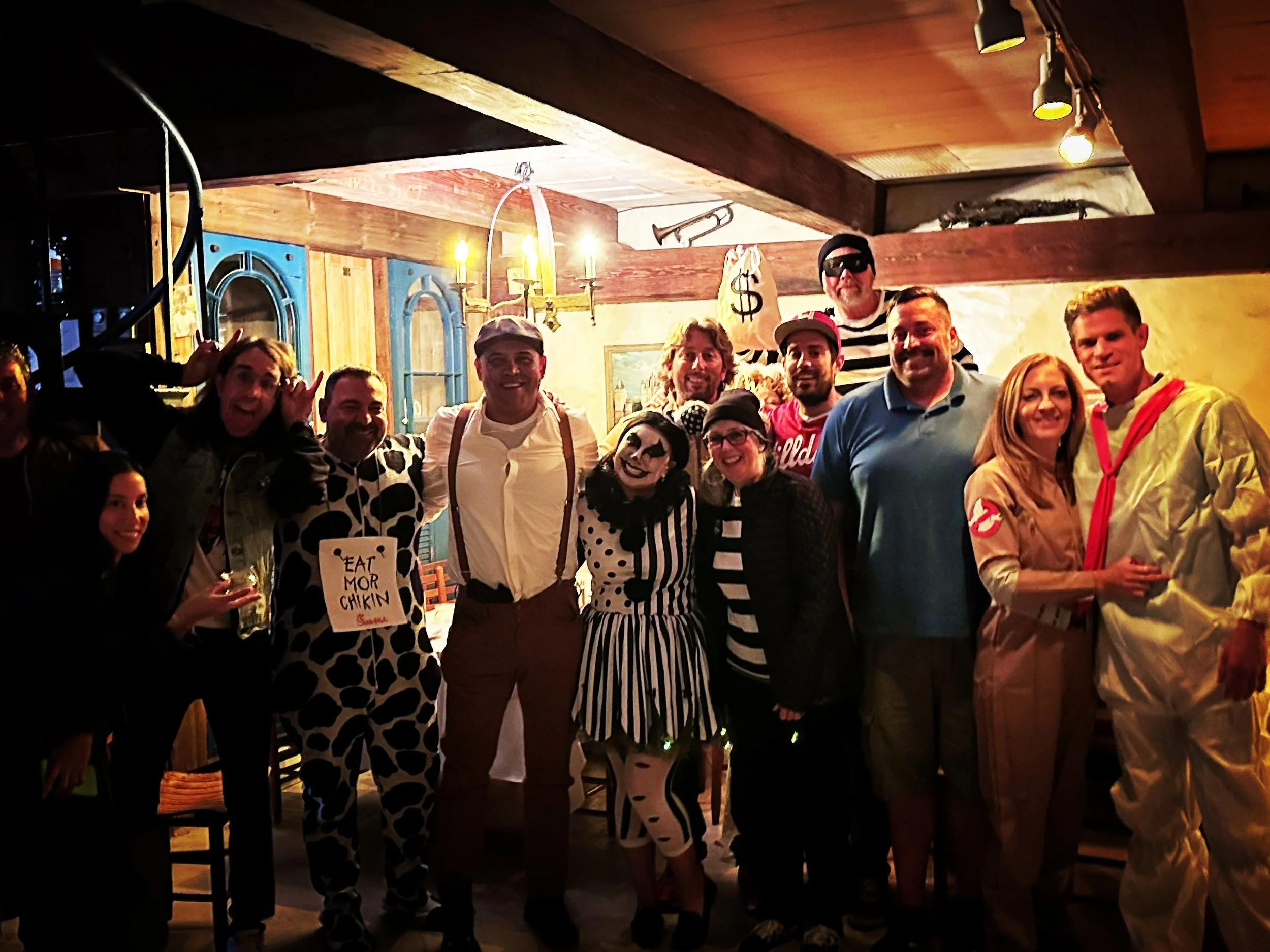 A group of people dressed up in costumes pose together in a dimly lit room decorated with musical instruments and vintage decor. Some are smiling and appear to be enjoying a themed party, with one person dressed as a clown and another wearing a Ghostbusters outfit. A person in the back is holding a bag with a dollar sign on it.