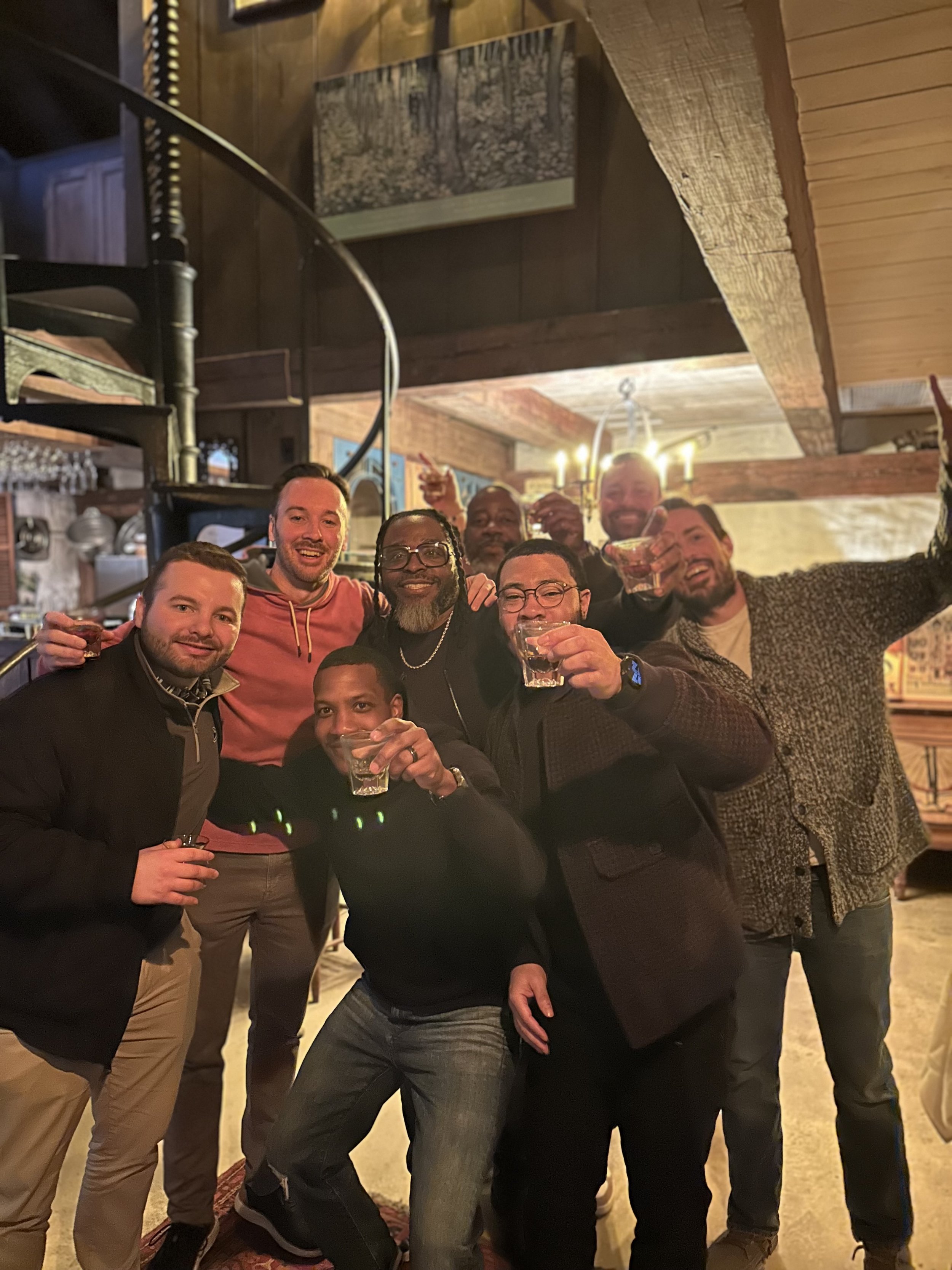 A group of men celebrating indoors, holding drinks, smiling, with a rustic wooden interior and a painting of a forest on the wall.