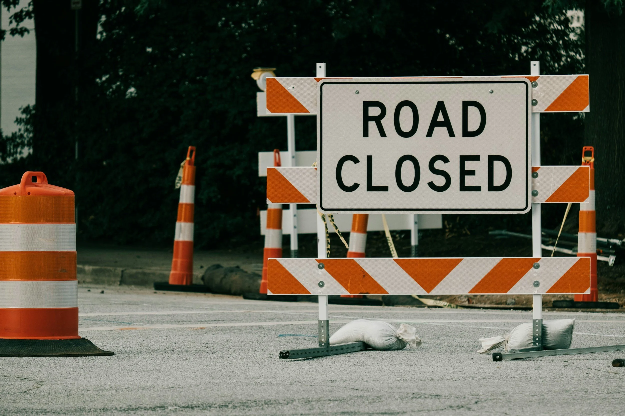 A street closed sign with orange and white barricades, orange traffic barrels, and sandbags blocking the road, with bushes and trees in the background.