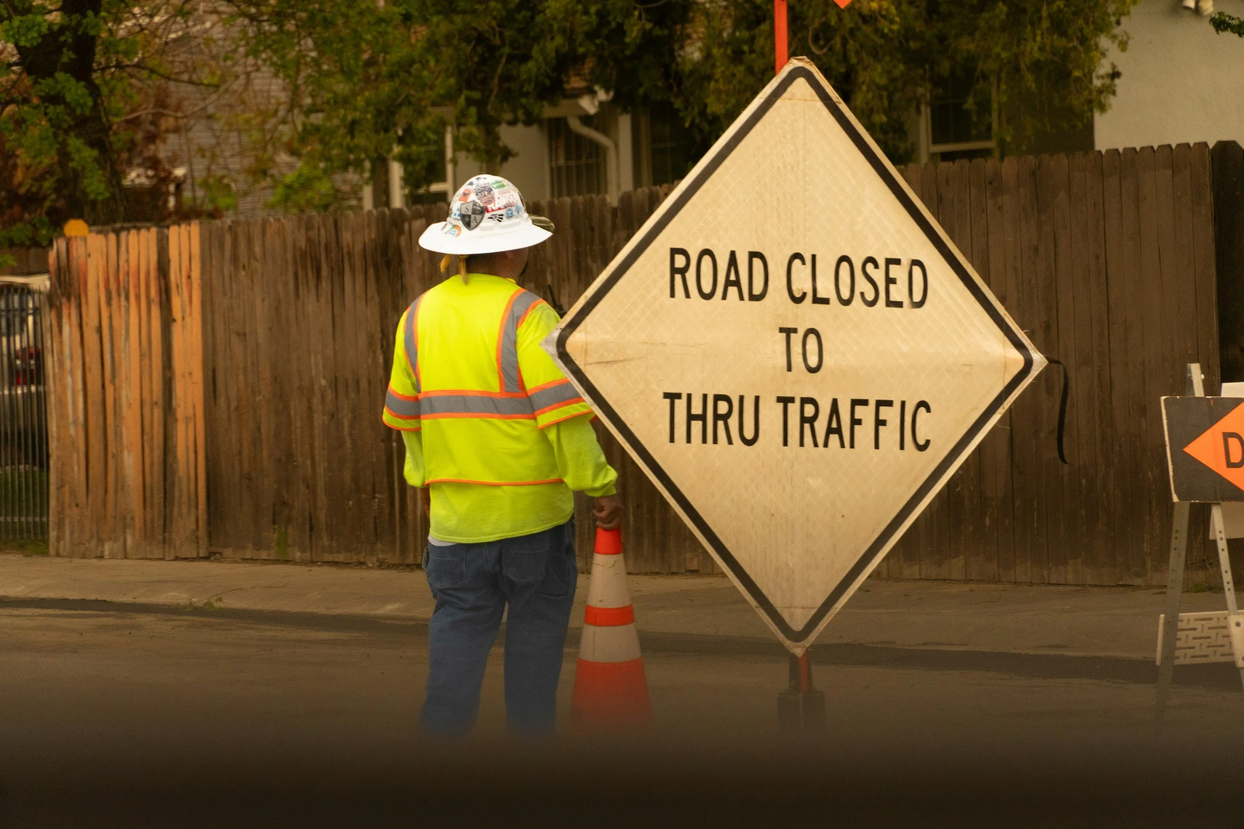 A construction worker wearing a high-visibility yellow safety jacket and a white hard hat with stickers, is standing near a large road closed sign and an orange traffic cone. The worker has their back to the camera and is facing a wooden fence and trees.