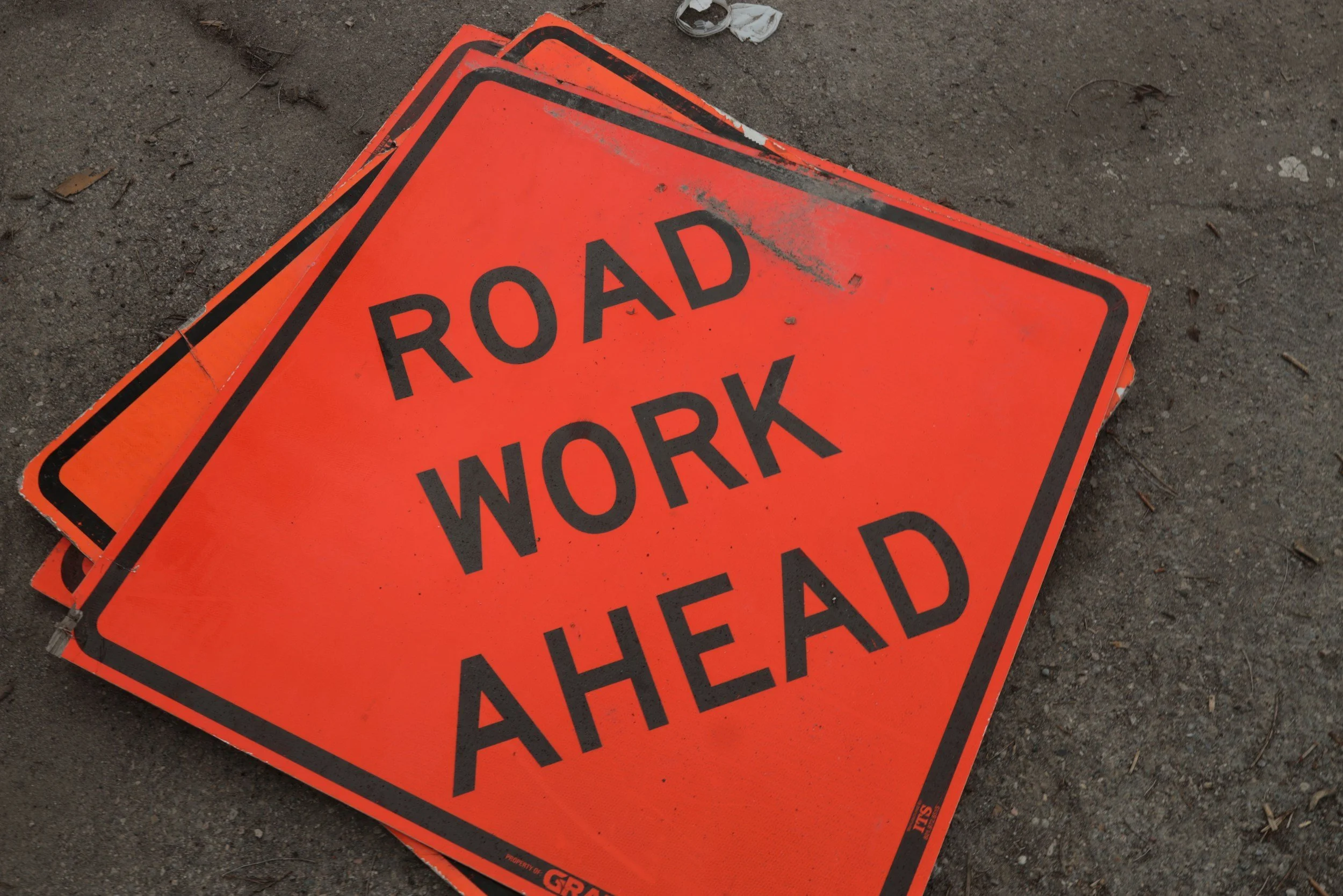 Several orange construction signs with black borders and text that reads 'ROAD WORK AHEAD' are lying on the ground on a dirt surface, with some small debris and a piece of white cloth nearby.