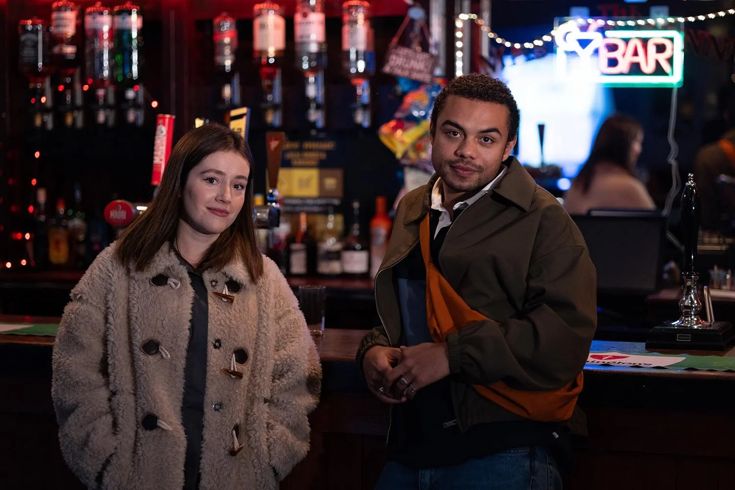 Zooport production still. Two people pose at a dimly lit bar with liquor bottles, taps, and a neon bar sign in the background.