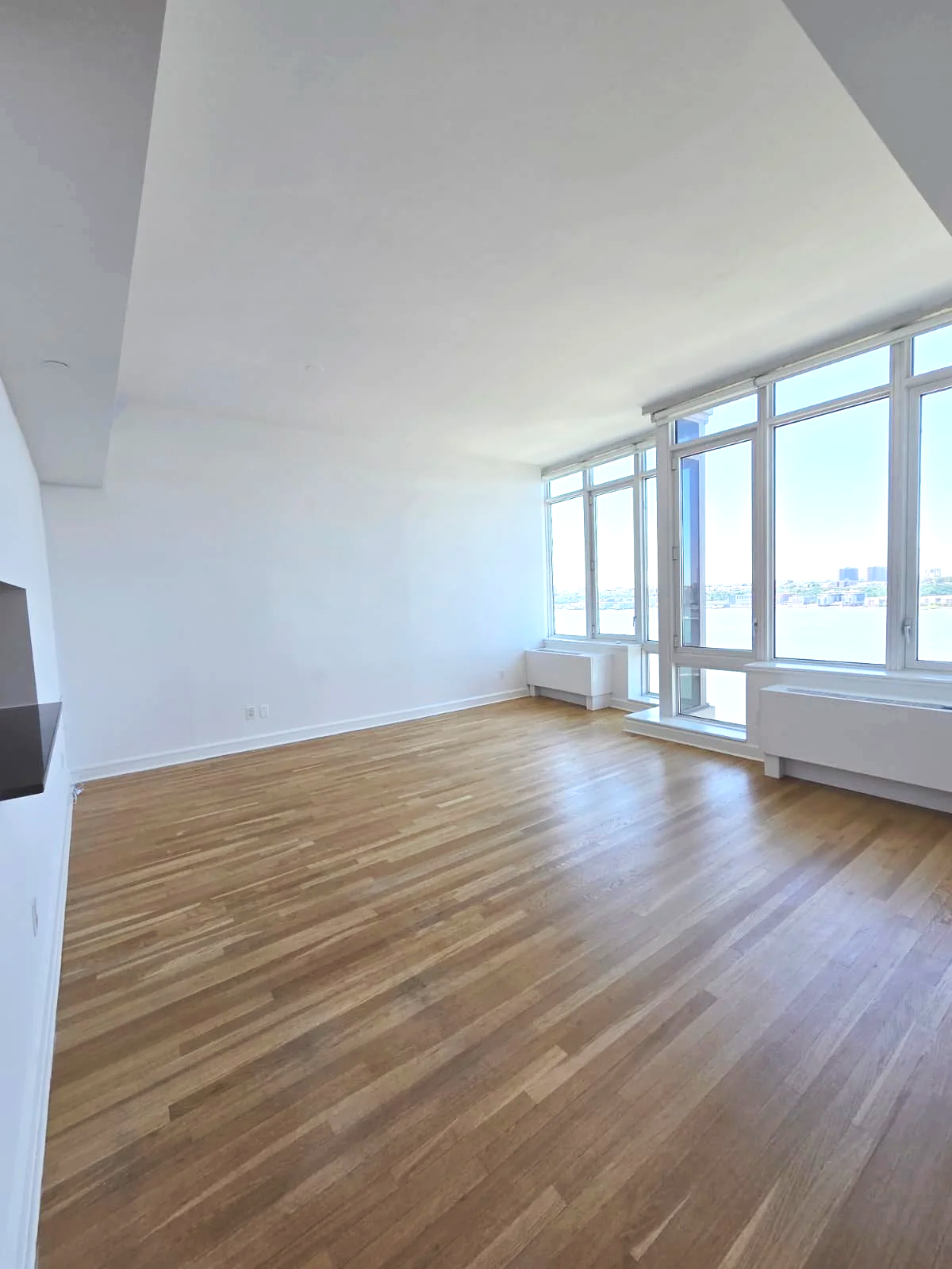 Empty living room with large windows showing a city skyline and water view, light wood flooring, white walls, and high ceiling.