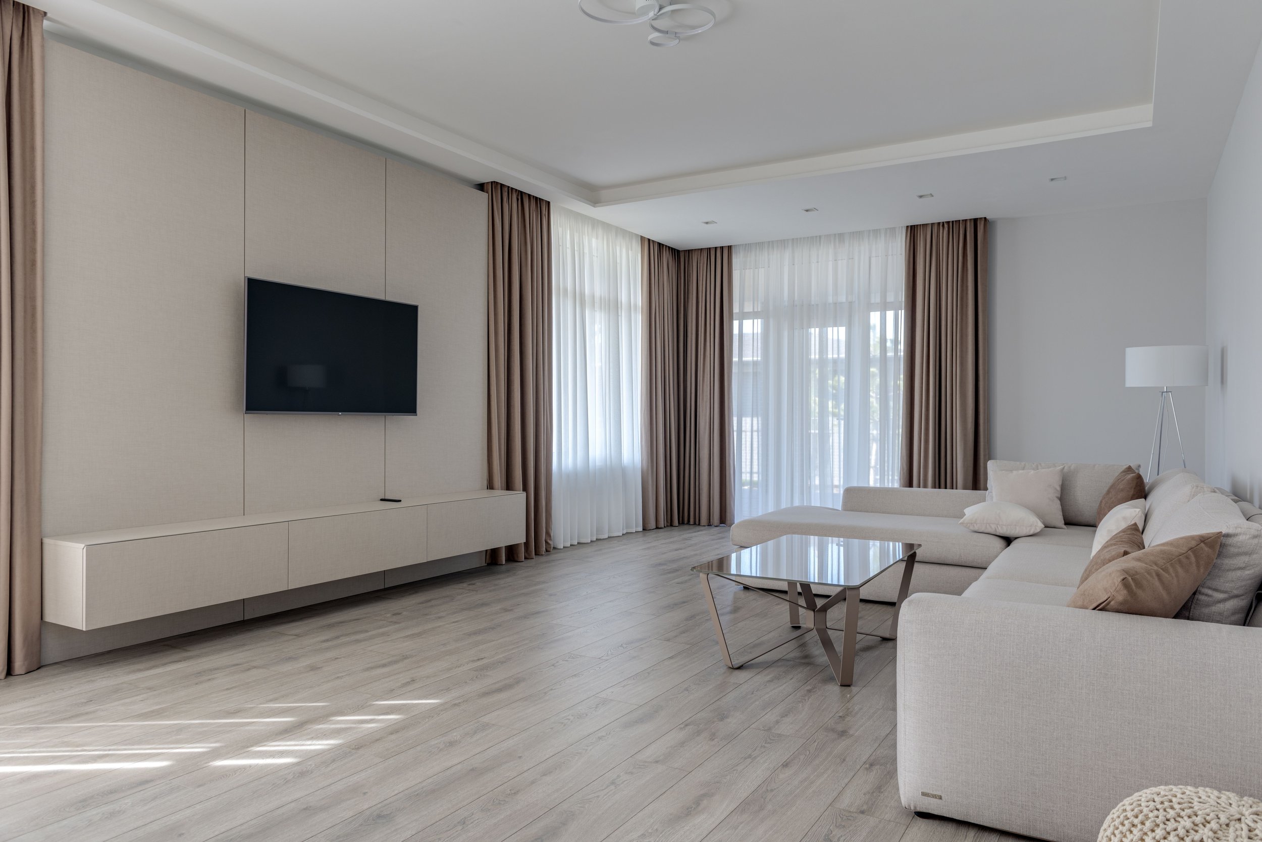 Bright living room with large windows and beige curtains, white sofa with pillows, and a modern coffee table. Flat-screen TV mounted on the wall and a floor lamp