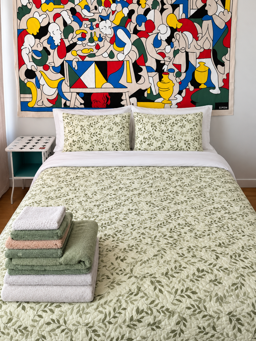 A neatly made bed with a green leafy patterned bedspread and matching pillows, topped with a stack of folded towels in white, beige, and green. Behind the bed, there is a colorful abstract painting hanging on the wall and a small white bedside table with black symbols or spots on its surface.