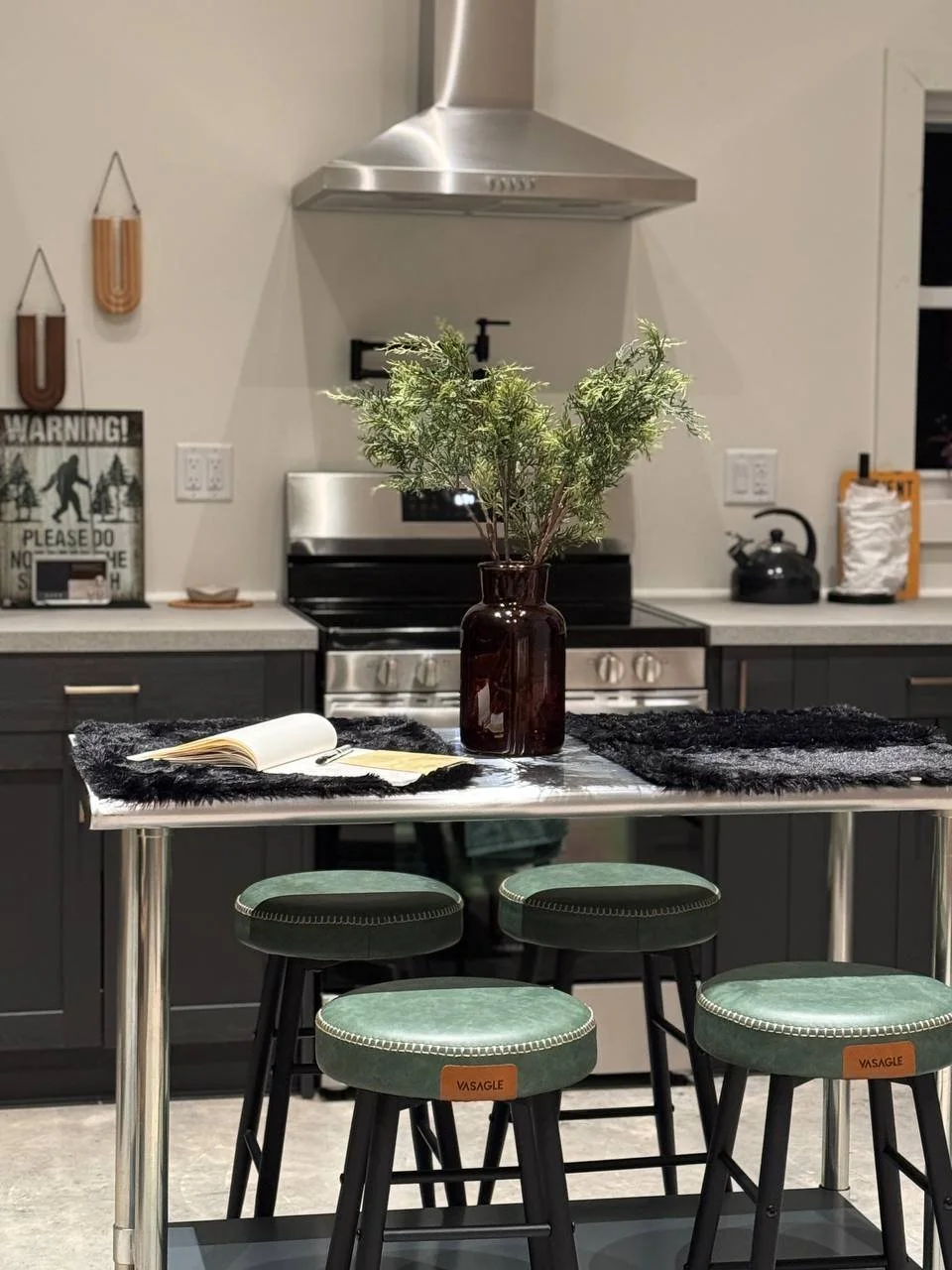 Modern kitchen island with greenery, an open book, black mats, green stools, dark cabinetry, and a stainless steel range hood.