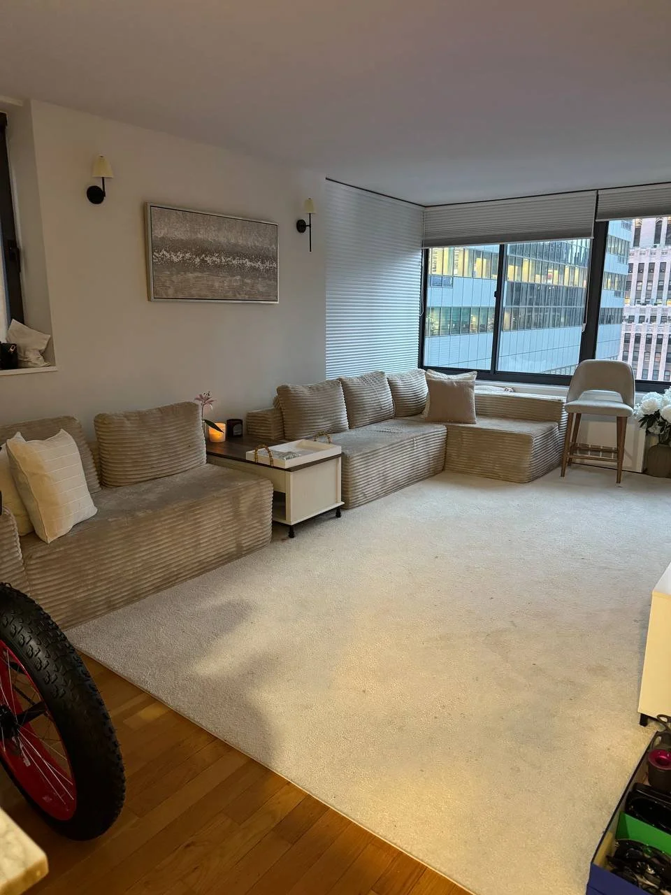 Bright living room with beige striped sofa, white side table, large window with city view, and white carpet.