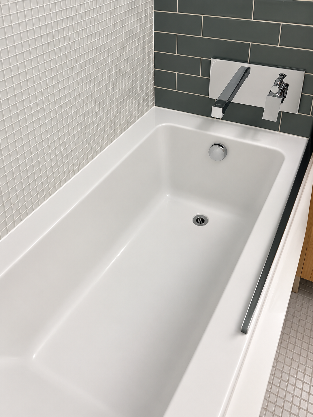 Modern white bathtub with a chrome faucet against tiled wall with small gray mosaic tiles and larger dark gray subway tiles.
