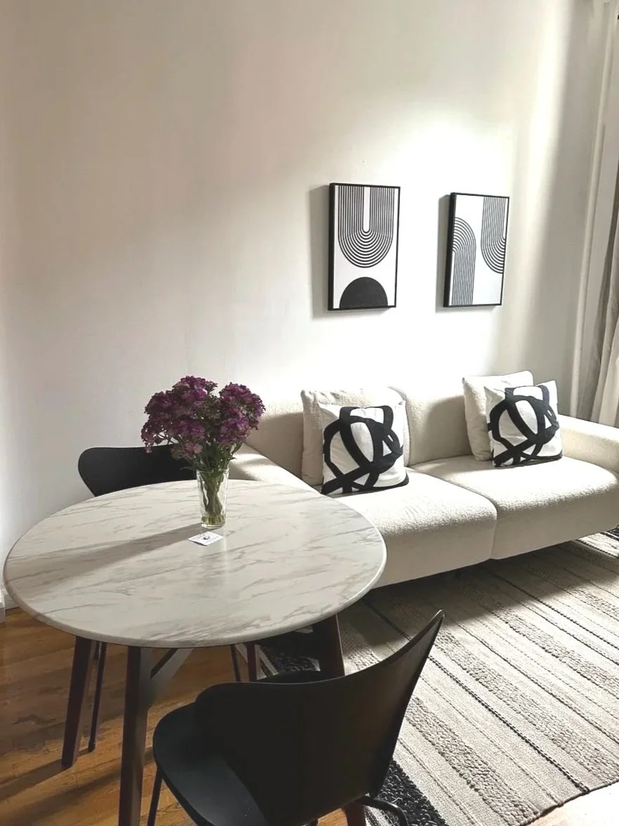 Living room with a white sofa, black and white decorative pillows, black and white abstract art on the wall, a round marble table with a vase of purple flowers, and a black chair, with a striped rug on hardwood floor.
