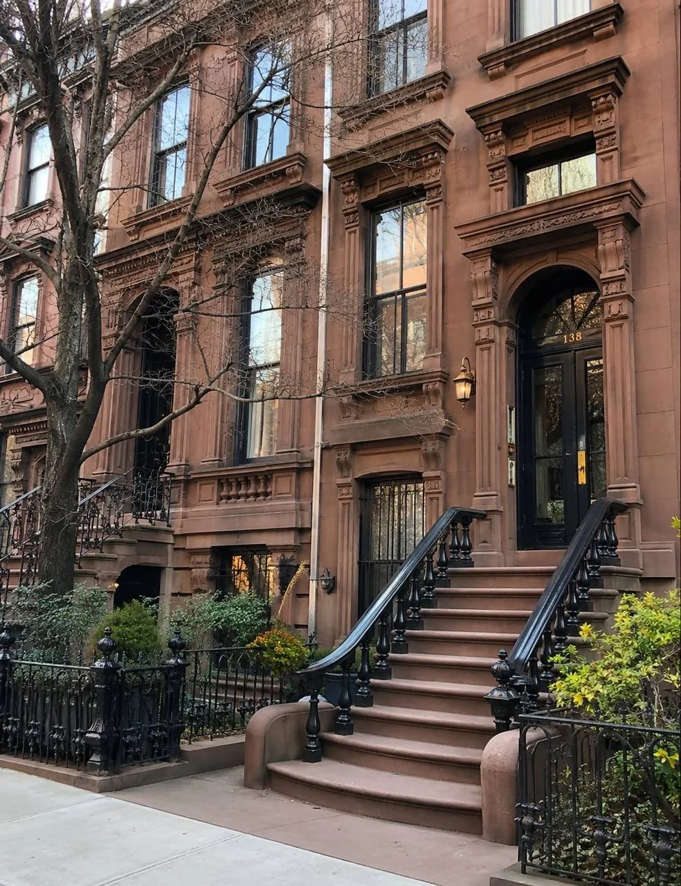 Brownstone entrance with stone steps, a black front door, iron railings, decorative window frames, a tree, and a small garden.
