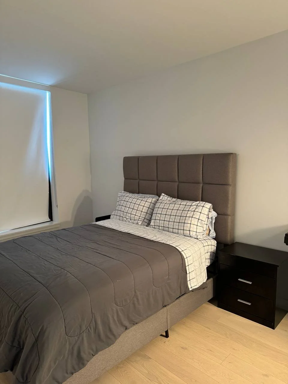 A neatly made bed with a brown quilt, checkered pillows, and a tall beige headboard in a minimalistic bedroom with light-colored walls and wooden floors, next to a black nightstand.