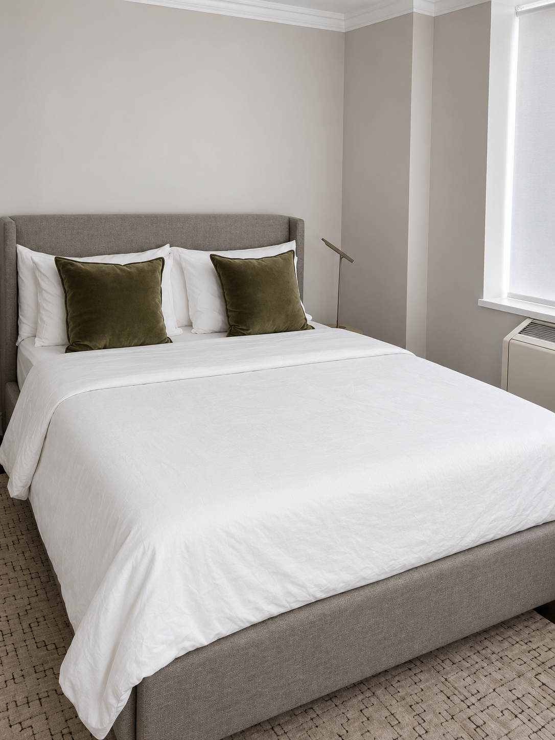 A neatly made bed with a grey upholstered headboard, white sheets, and two green velvet pillows in a bedroom with white walls and a window.