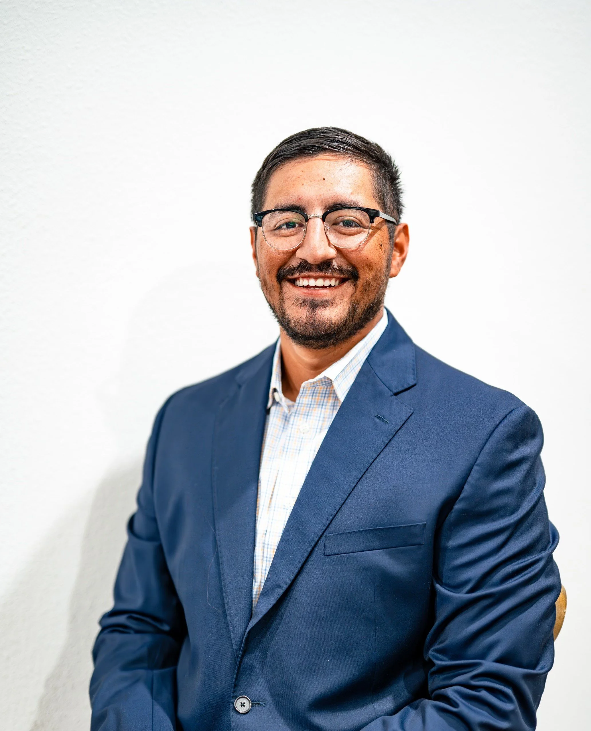 Portrait of a smiling man wearing glasses, a navy blue suit, and a checkered shirt, sitting against a plain white background.