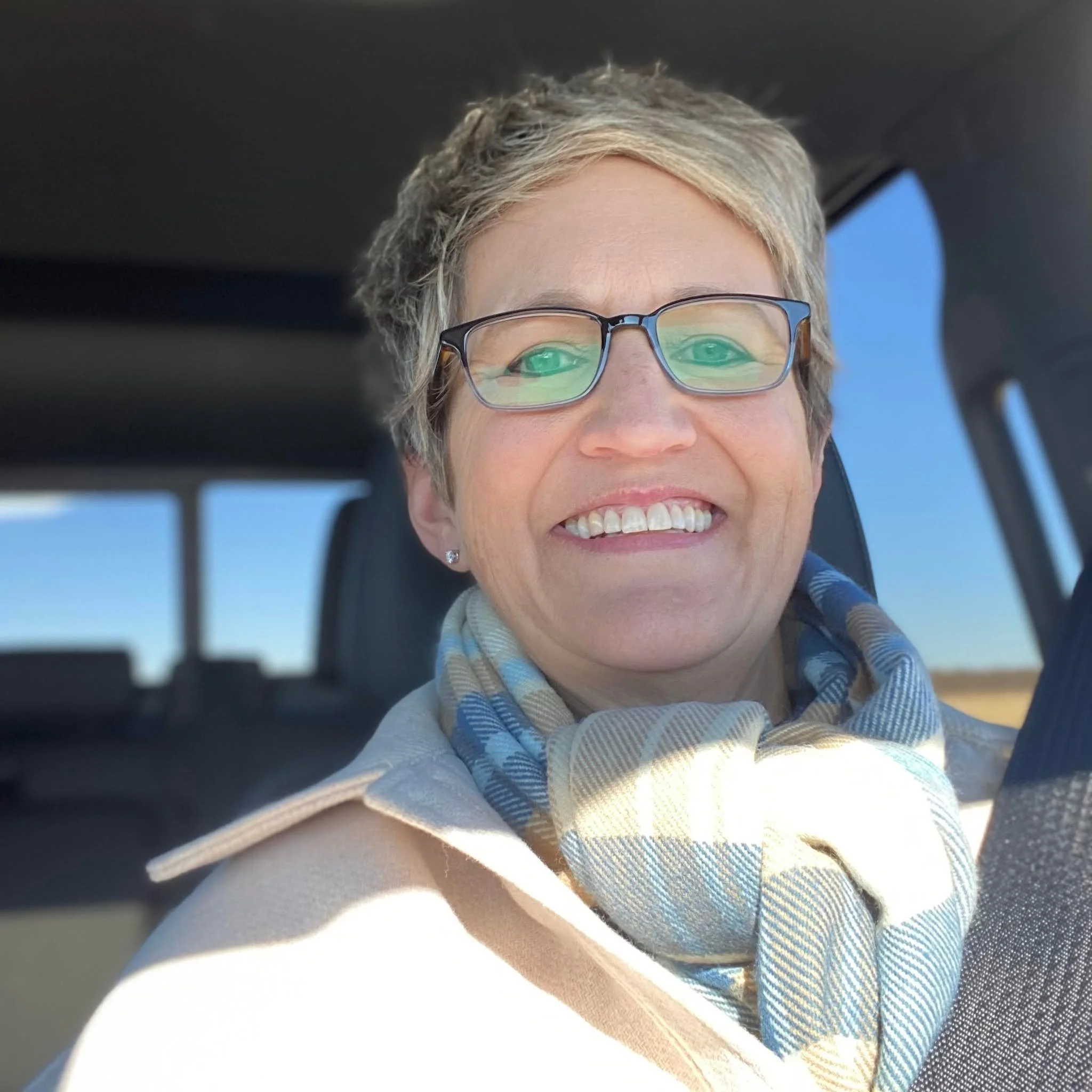 A smiling person wearing glasses with a brief glimpse of their face, in an environment with a bright sky visible through a window or opening.