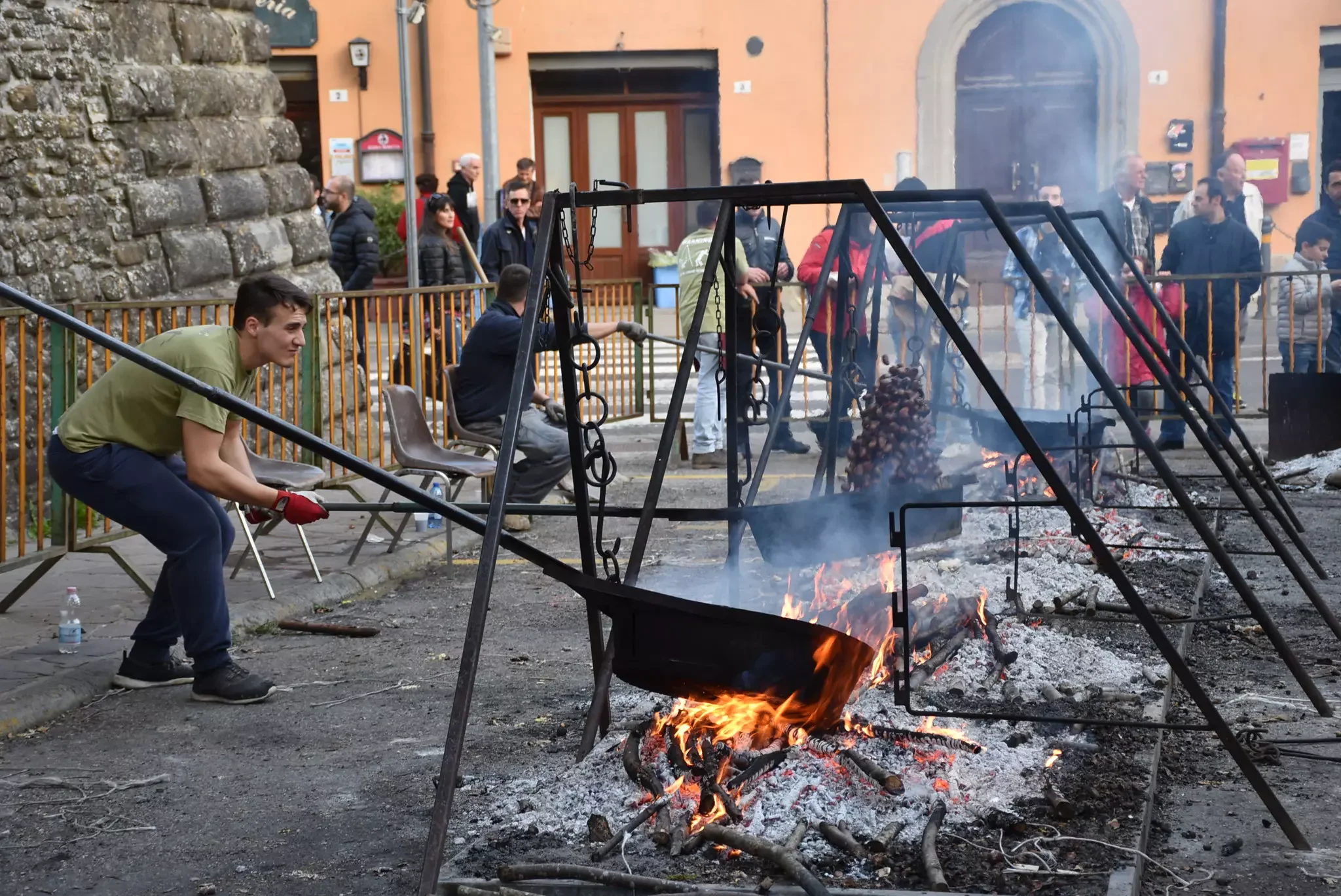 Gruppo di persone intorno a un falò in una piazza, alcuni con mascherine, in un contesto urbano.