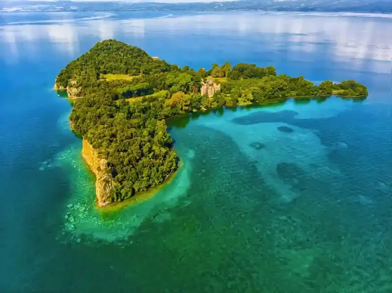 Isola verde ricoperta di alberi circondata da acque cristalline e limpide, con alcune parti di fondale visibile, vista aerea.