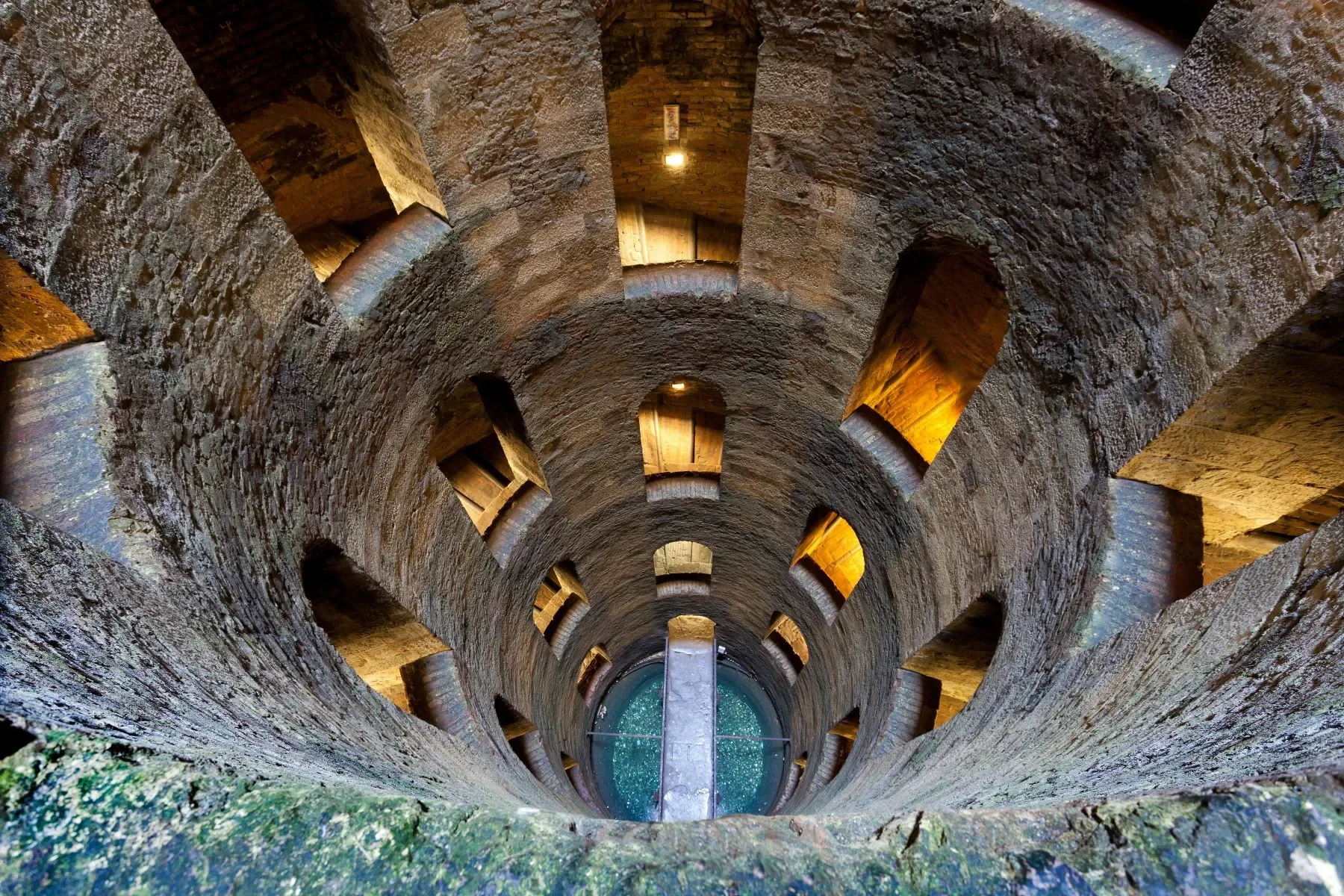 Vista dall'alto di un pozzo o silos con pareti di pietra e finestre rettangolari, con luce calda proveniente dall'esterno